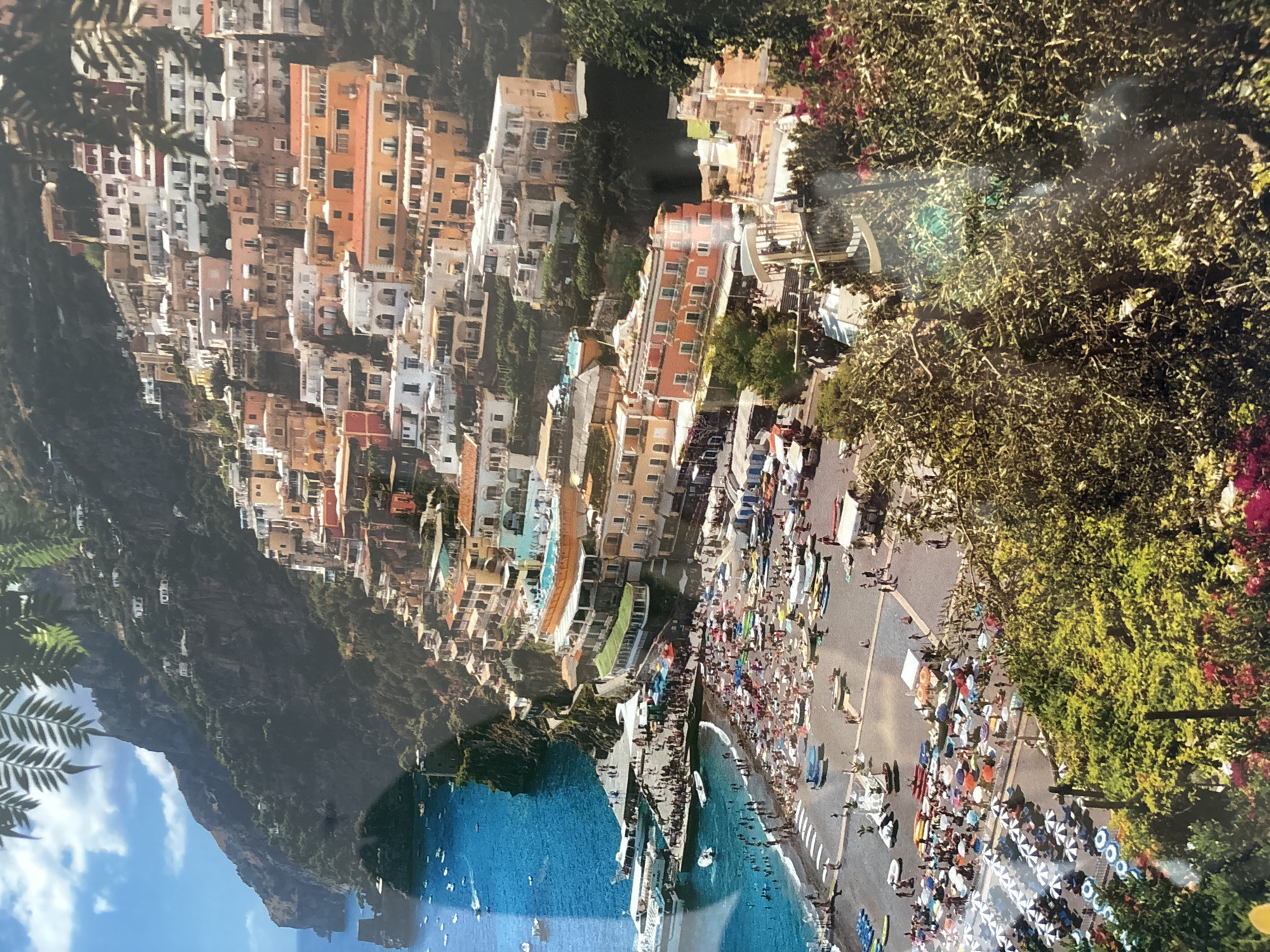 This is a photograph of Positano, Italy, a coastal town in the province of Salerno, Campania, southern Italy. The image depicts a vibrant, colorful village built into a steep cliffside overlooking the turquoise waters of the Mediterranean Sea. Buildings are painted in shades of yellow, orange, pink, and white, with terracotta roofs. They cascade down the hillside, interspersed with lush green vegetation.
In the foreground, a bustling beach is filled with numerous beach umbrellas and sunbathers. Loungers are arranged in neat rows, and people are swimming in the sea. Boats are anchored offshore and docked at a pier where crowds are gathered. Several ferries and smaller vessels are visible in the water.
The cliff face is covered in dense greenery, with a few scattered buildings nestled among the trees. The sky is a bright blue with some scattered white clouds. The overall impression is one of a lively and picturesque tourist destination on a sunny day. There is no indication of this location being in Dubai, United Arab Emirates.