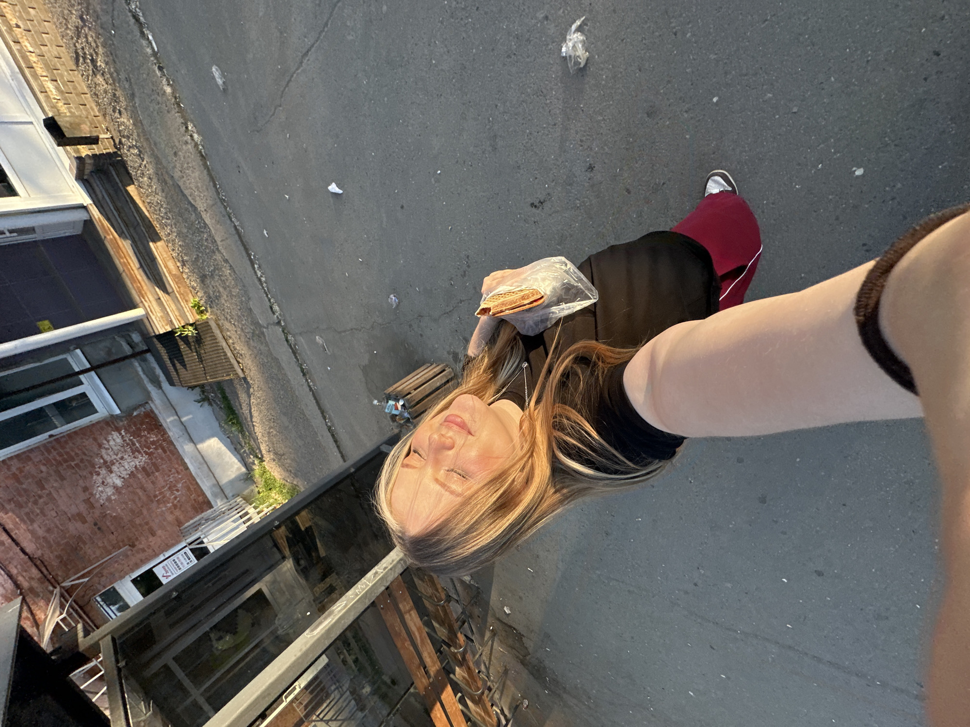 A young woman is holding up her cell phone, likely to take a selfie in an urban area, with the camera oriented at an angle. She is holding a wrapped food item in her left hand and wearing a black top and red pants with white stripes. The setting appears to be an alleyway or side street, with a building to the left and a concrete or asphalt ground. Some trash is scattered on the ground. Based on prior knowledge, the location of Chaikovsky, Russia is unknown.