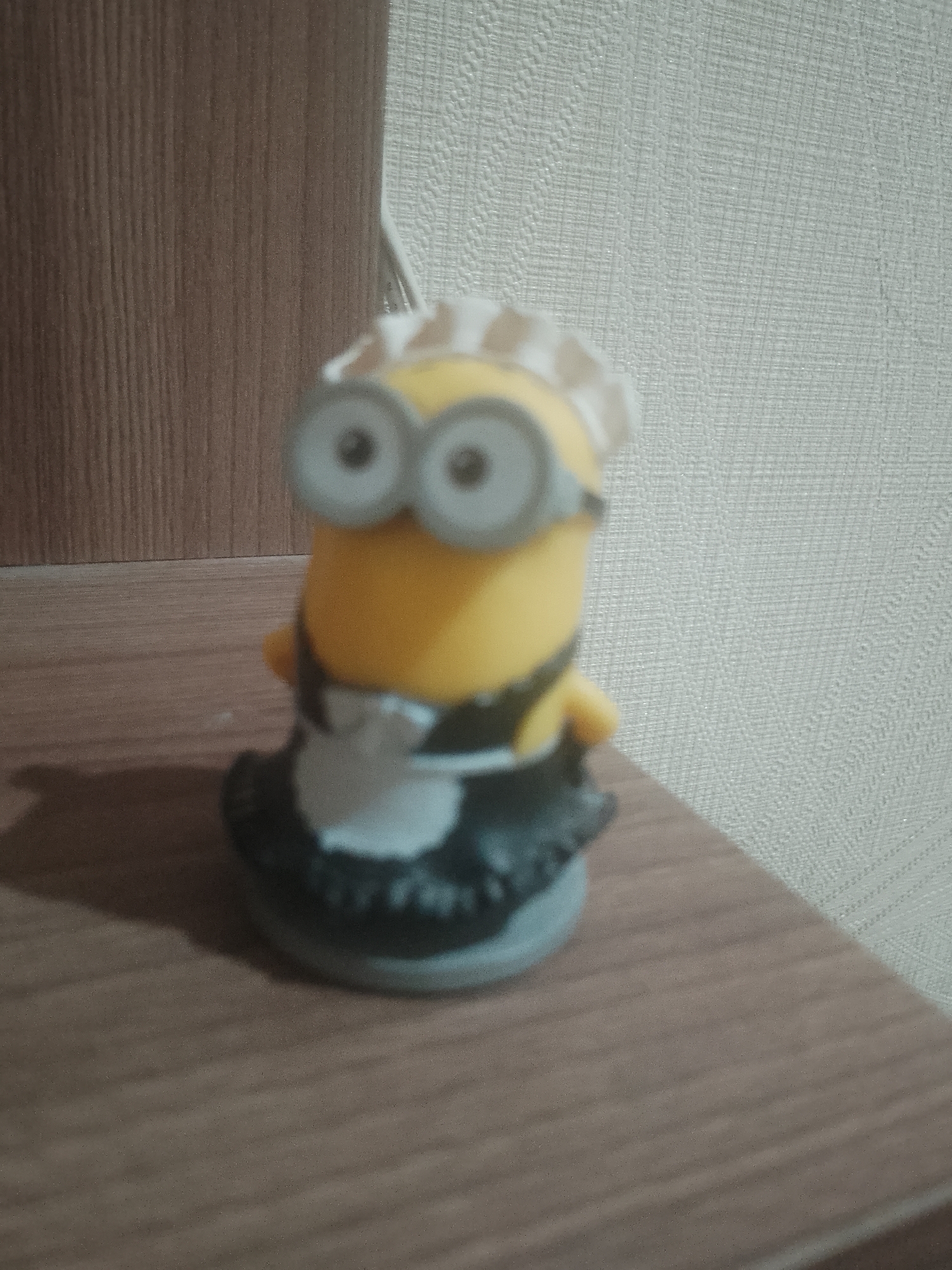 The image shows a plastic toy Minion figure dressed as a maid. The figure is yellow with gray goggles and is wearing a white frilly cap, a white bib, and a black dress with a frilly edge. It stands on a brown, wood-grain surface. A light-colored textured wall is in the background, and a dark brown wood panel is on the left. The location is not discernable, and there is no information indicating the location is in Noviy, Russia.