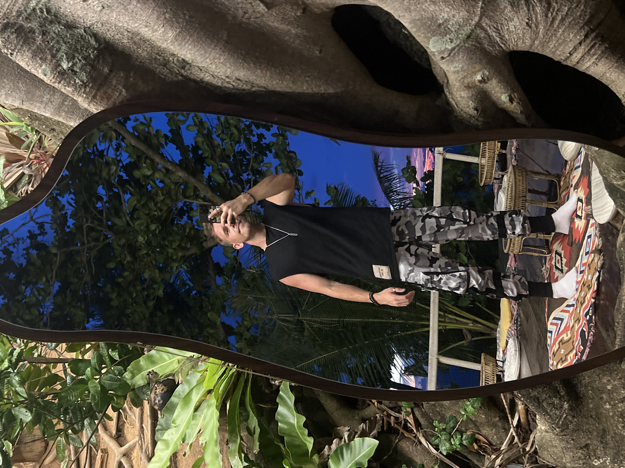 The image is a mirror selfie taken by a young man standing in what appears to be an outdoor setting. The man is wearing a black t-shirt and camouflage pants. He is holding a phone in his right hand, angled to capture his reflection in the mirror. The mirror itself has an irregular, organic shape and is framed by the gnarled roots of a large tree.
Behind the man in the reflection, a tropical scene unfolds. Lush green foliage, including palm fronds, is visible, suggesting a warm climate. In the background, there are glimpses of woven chairs and tables, and a patterned rug or blanket, indicating an outdoor seating area. The sky is a deep blue, transitioning to lighter shades of purple and pink near the horizon, suggesting either sunrise or sunset.
The overall mood is relaxed and casual. The lighting indicates it is either twilight or early evening. The presence of abundant plant life and the style of the furniture suggest a tropical resort or a restaurant with an outdoor garden area. The image is taken in Karon, Thailand, which is consistent with the tropical vegetation. No text is clearly visible in the image.