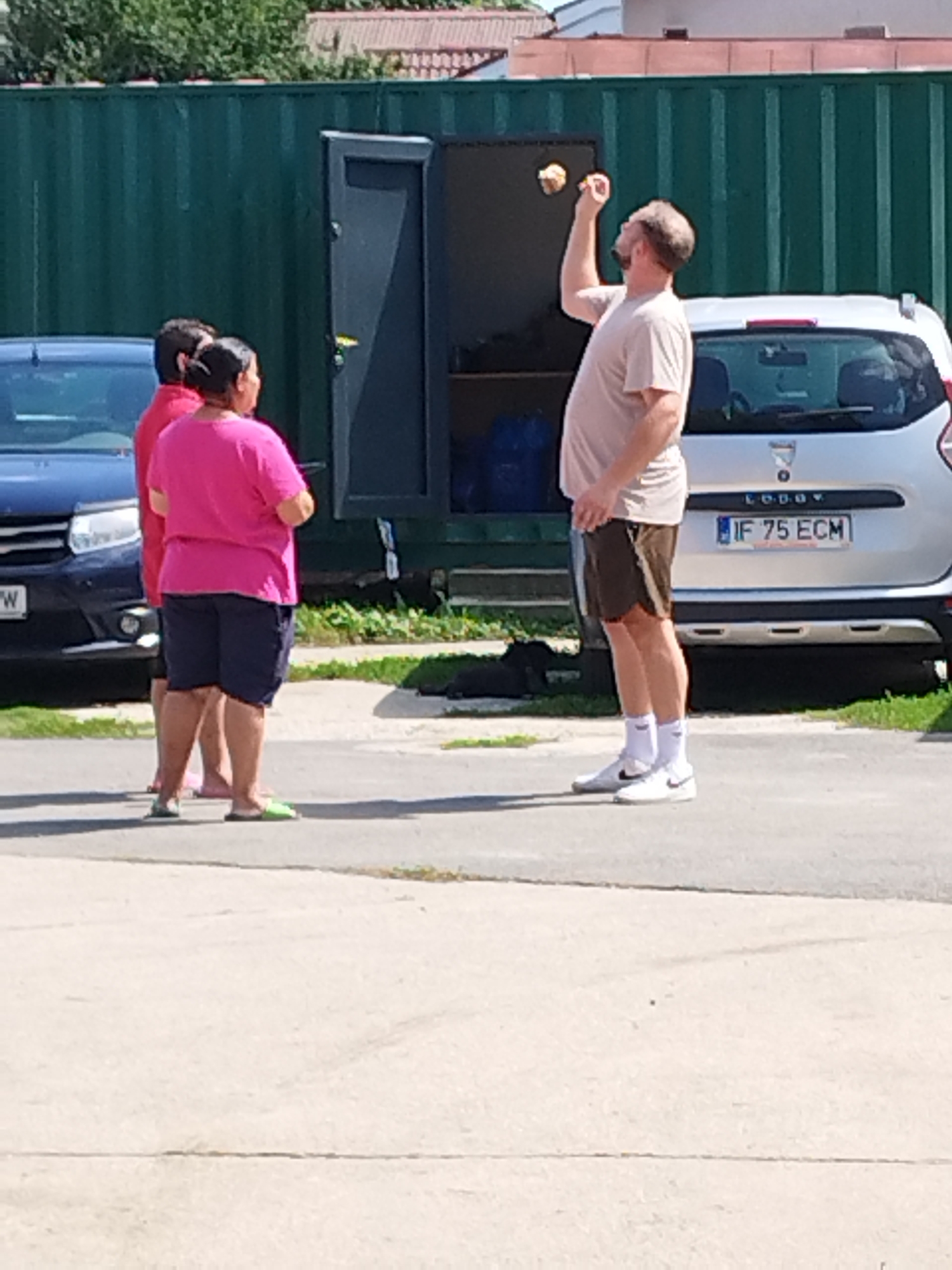 In Bucharest, Romania, on a sunny day, three individuals are visible in front of a large green industrial container. A man in a light-colored t-shirt and brown shorts is throwing two round objects, possibly apples, into the air. He is wearing white socks and white sneakers. Beside him, a woman in a bright pink t-shirt and dark shorts, with her hair pulled back, stands with her hands clasped. Behind her, a person in a red t-shirt is partially visible. Two cars are parked nearby: a dark blue sedan to the left and a silver hatchback with the license plate "IF 75 ECM" to the right, parked in front of the container. The ground is paved with concrete and asphalt.