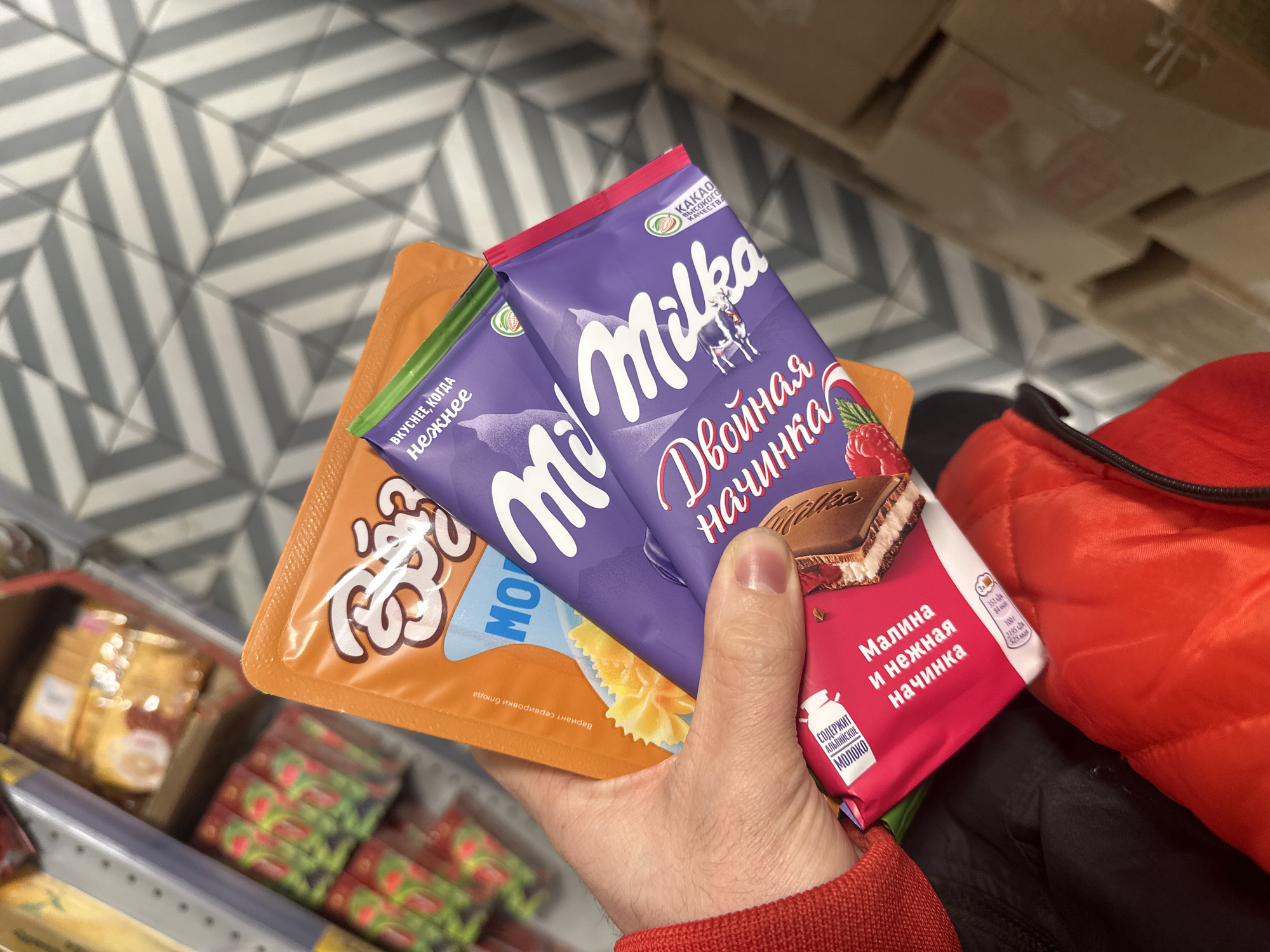 A hand holds an orange package of pasta next to two purple Milka chocolate bars with Russian text that translates to "double filling" and "raspberry and soft filling." The hand is wearing a red sleeve, and a red jacket is visible in the background. The products appear to be on a shelf in a store with a chevron pattern floor. The items are likely located in Moscow, Russia, given the Russian language on the packaging.