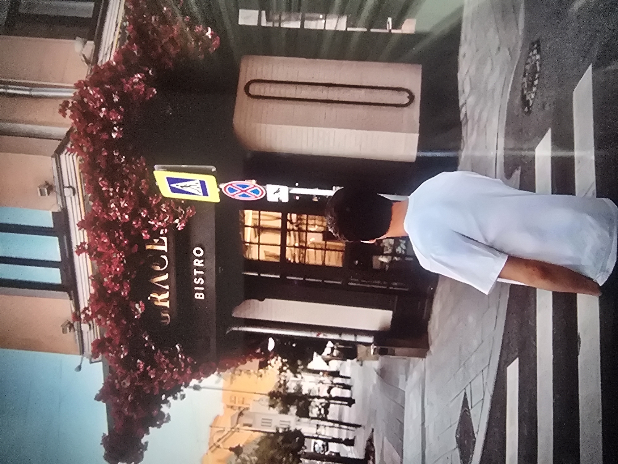 The image captures a street scene in a city, with a man in a white t-shirt standing on a crosswalk. The man is facing away from the camera, looking towards a building adorned with red flowers. Above the entrance of the building, the word "GRACE" is visible in large, ornate lettering, followed by "BISTRO" in smaller text. A yellow pedestrian crossing sign with a blue triangle is mounted on a pole to the left of the entrance. The building appears to be a restaurant or cafe. The street is paved with cobblestones, and trees line the sidewalk. The overall atmosphere is urban and somewhat picturesque due to the floral decoration.