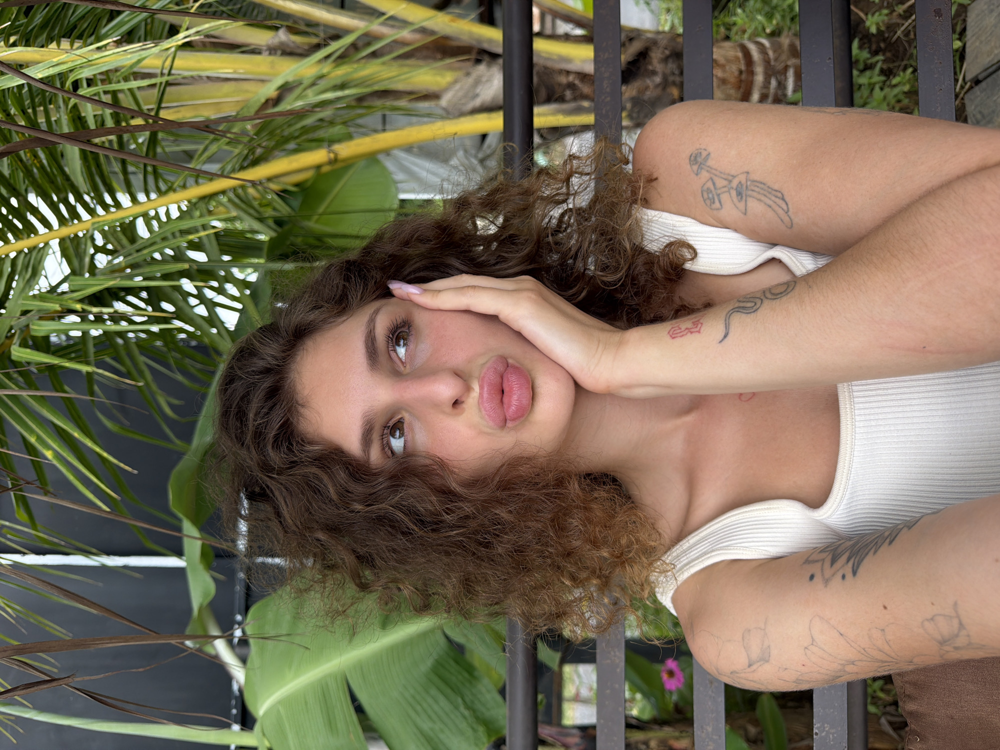 A young woman with curly brown hair, wearing a white ribbed tank top, poses for a photo with her hand on her cheek and her lips pursed. She has tattoos on her arms, including what appears to be flowers on her left arm and mushrooms on her right. She is seated outdoors, with lush green foliage and palm trees in the background. The scene is casual and relaxed, suggesting a warm, tropical environment.