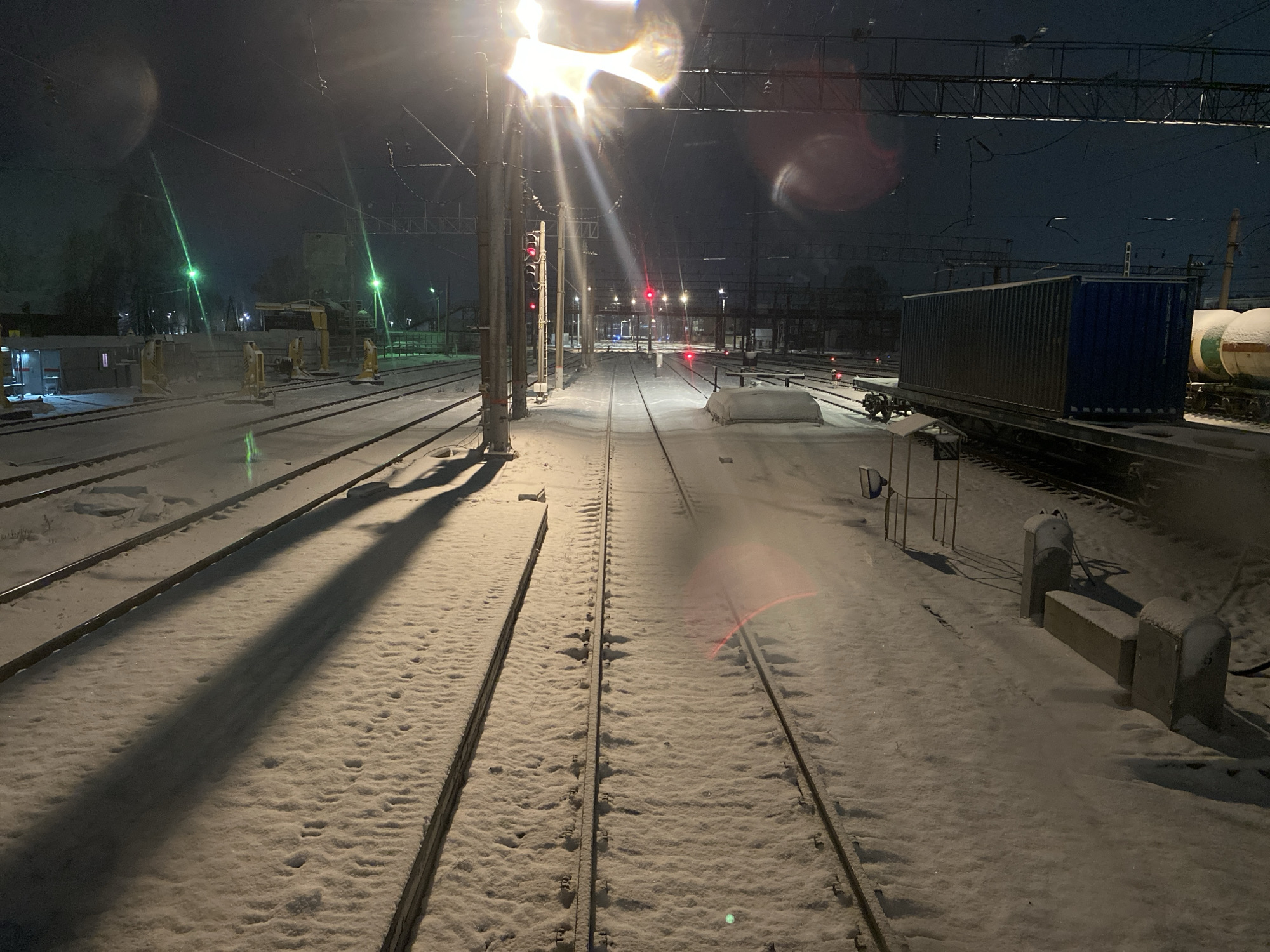 The image depicts a snow-covered railway yard at night in what is assumed to be Agryz, Russia. Multiple sets of parallel train tracks are visible, stretching into the distance under dim lighting. To the right of the image, there is a train with a blue container and a tank car. There are some auxiliary structures, such as utility poles, lights, and other rail yard equipment. The image is taken at ground level and the angle is slightly upward. There is snow covering the ground. In the distance, there are lights and buildings suggesting a city environment. The lighting conditions are low and harsh. The overall scene evokes a cold, industrial atmosphere.