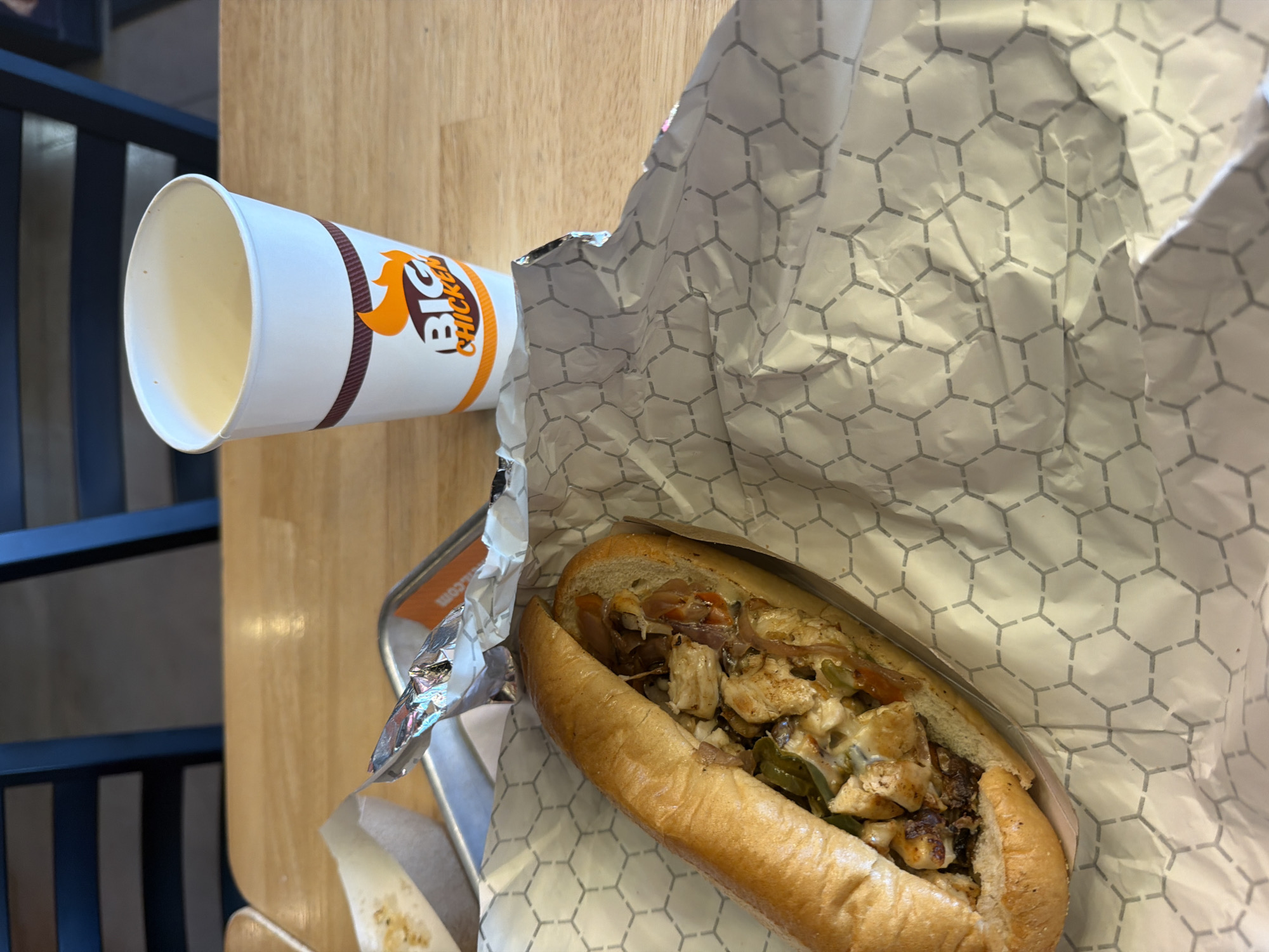 The image captures a meal setting. The meal consists of a sandwich filled with what appears to be grilled chicken, onions, and green peppers, possibly with cheese. The sandwich is placed on a patterned paper wrapper. Next to it is a white paper cup with the "BIG CHICKEN" logo in orange lettering. The setting is on a light wooden surface, and part of a dark chair is visible in the background. Given the presence of the "BIG CHICKEN" branding, it is likely that this scene takes place at a Big Chicken restaurant location. The restaurant is most likely in Rosemont, United States of America, given that it is the only location with a Big Chicken restaurant.
