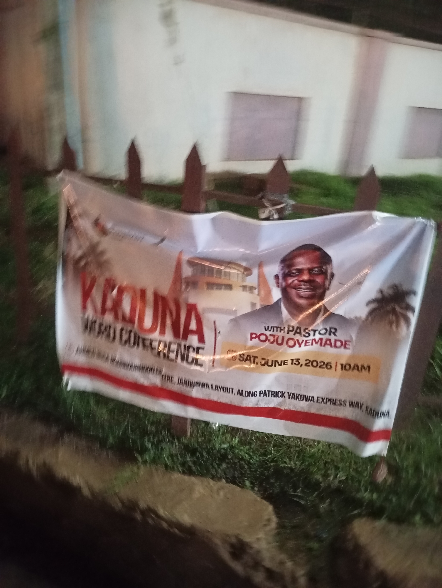 The image depicts an outdoor scene in the city of Unknown, Unknown, primarily featuring a white banner advertising a religious event. The banner, oriented horizontally, displays in prominent red text "KADUNA WORD CONFERENCE." A central image on the banner is a smiling, bald man, identified by the text "WITH PASTOR POJU OVEMADE," wearing a light-colored suit jacket over a collared shirt. Below his image, the event date and time are printed: "SAT, JUNE 13, 2026 | 10AM." Further down, an address or location is partially visible, reading "THE. JANJ... LAYOUT, ALONG PATRICK YAKOWA EXPRESS WAY, KADUNA." The banner's design incorporates graphic elements of palm trees and what appears to be a multi-story building. A red stripe runs along the bottom edge of the banner. The banner is attached to a dark-brown wooden fence, characterized by multiple pointed pickets. In the immediate foreground, a patch of green grass is visible, transitioning into a dark, indistinct area below. In the blurred background, a light-colored building with at least two dark, rectangular openings, possibly windows or boarded sections, is discernible. The overall lighting suggests dusk or night, with artificial light illuminating the banner. The image exhibits some blur, particularly towards the edges and in the background.
