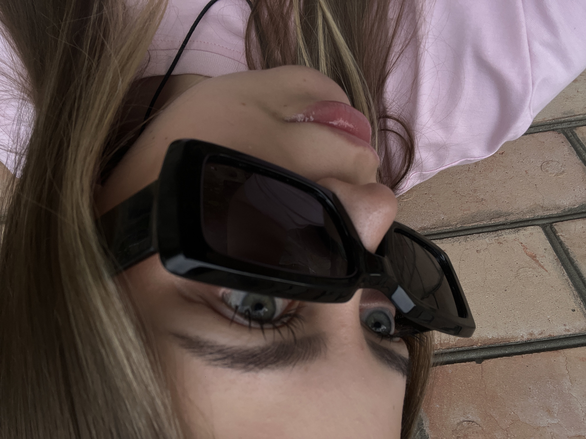 A young female with light skin, green eyes, and long blonde hair is shown from the chest up, looking to her left. She is wearing black-rimmed sunglasses and a pink t-shirt. The background consists of a brick wall. The image appears to be taken outdoors. No specific location within Novopokrovskaya, Russia, can be determined from the visual information provided.