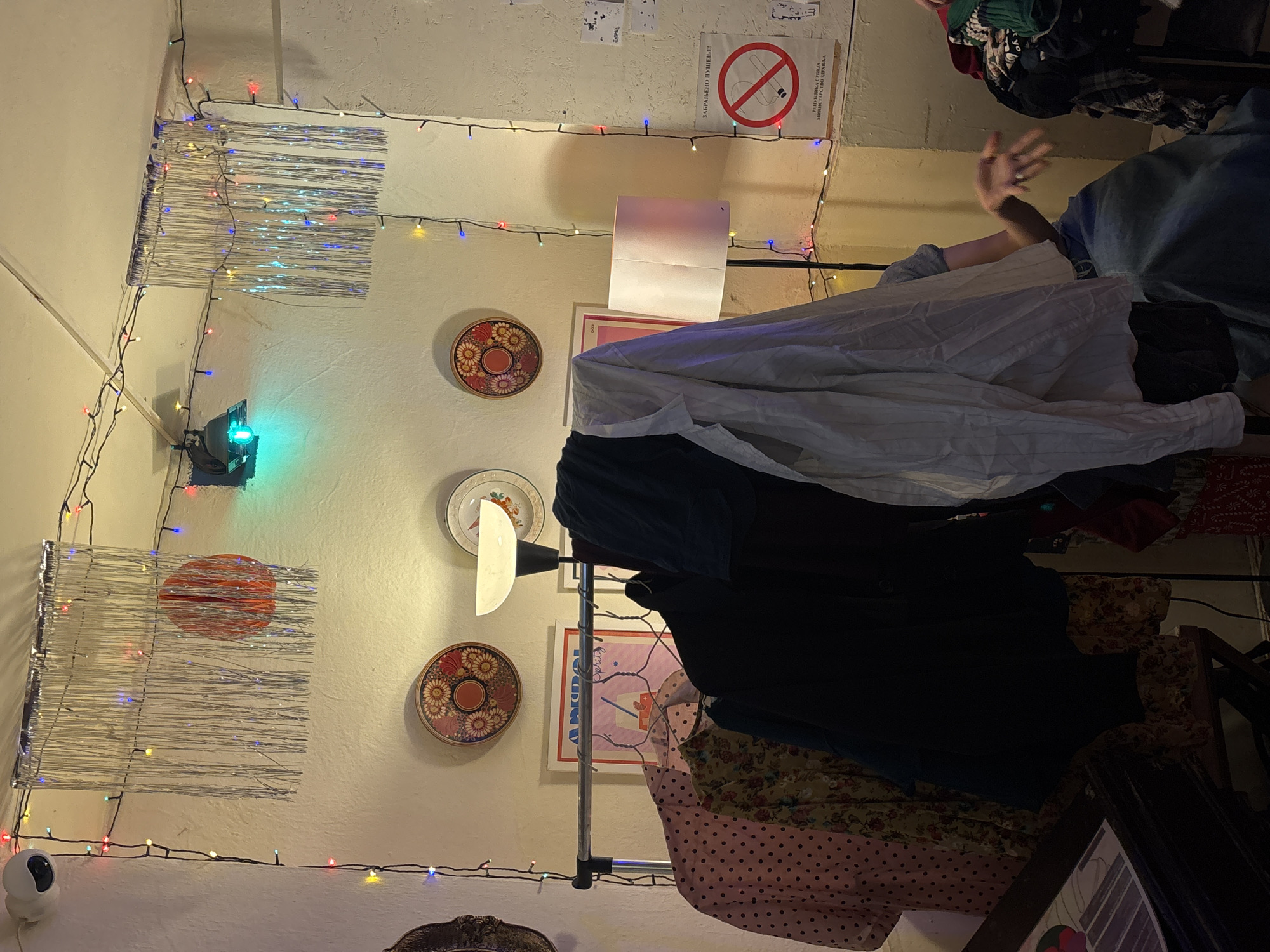 The image depicts a cluttered indoor space, possibly in Belgrade, Serbia, indicated by the Cyrillic script on a "No Smoking" sign on the wall. The room is decorated with strands of colored lights and silver foil decorations. There are several plates hanging on the wall. A clothing rack is heavily laden with clothes, covered with a white shirt or sheet. A person's hand is visible near the top right of the frame. A security camera is mounted in the lower-left corner of the frame. The room's lighting appears dim.