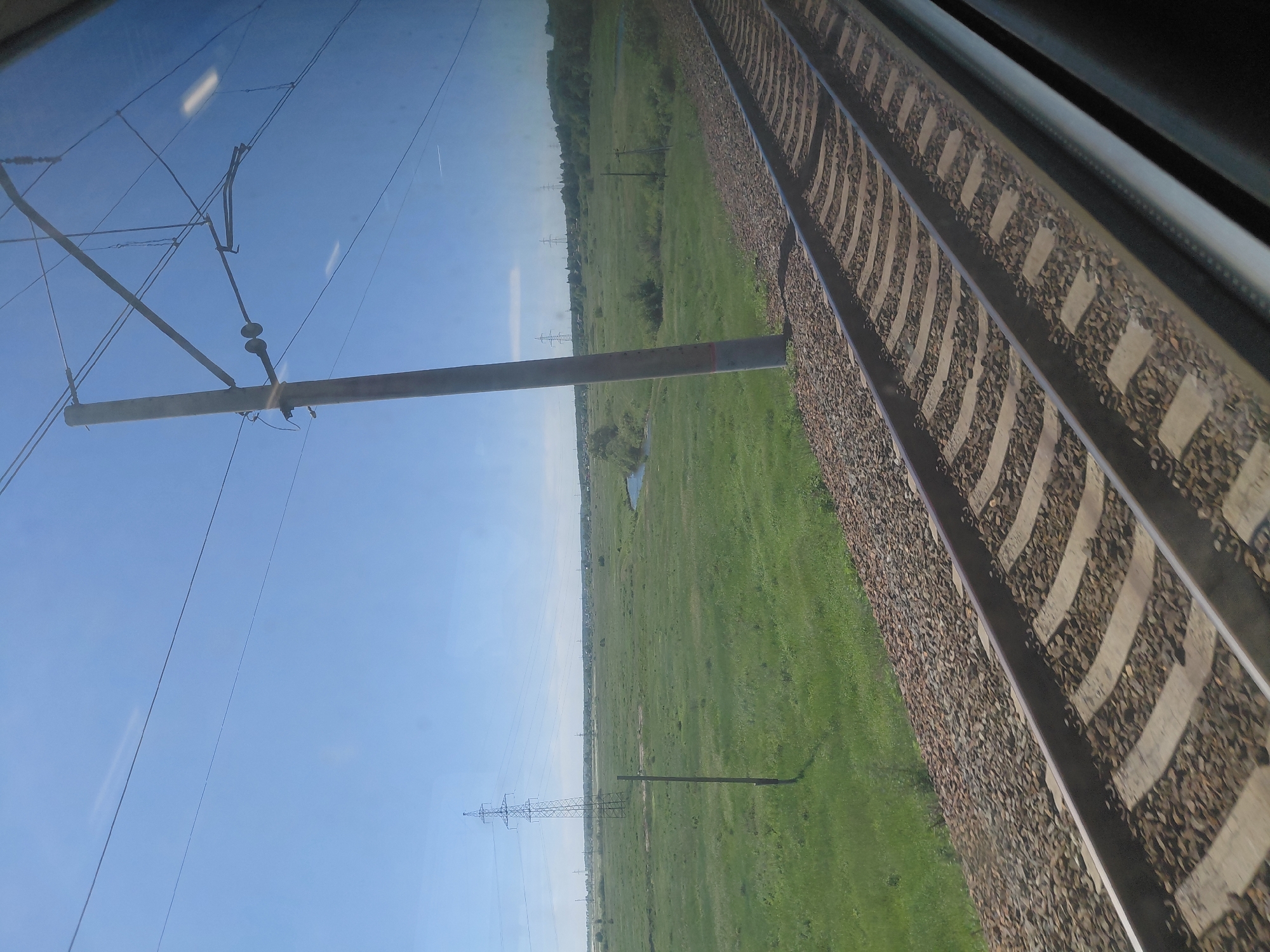 The image shows a view from a train window, looking out at a rural landscape. The foreground features a set of train tracks, with ballast and concrete sleepers visible. To the left of the tracks is a grassy field, dotted with a few small trees and a single wooden pole. In the middle ground, the field continues, with a narrow body of water and several power pylons visible in the distance. The background is dominated by a clear blue sky. The location is identified as Chik, Russia. There are no people or animals visible in the image.