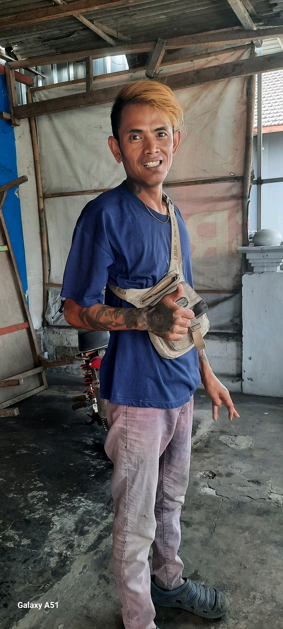 A full shot shows a man with bleached and styled hair standing in what appears to be a workshop or storage area. He is wearing a dark blue t-shirt and light-colored pants, with a beige crossbody bag slung over his shoulder. The man has tattoos on his arms and neck, and he is smiling at the camera while giving a thumbs-up gesture. In the background, a motorcycle is partially visible, along with various building materials and a corrugated metal wall. The floor is concrete and shows signs of wear and tear. The image also contains a watermark in the bottom left corner indicating "Galaxy A51". The location is identified as Selopuro, Indonesia.