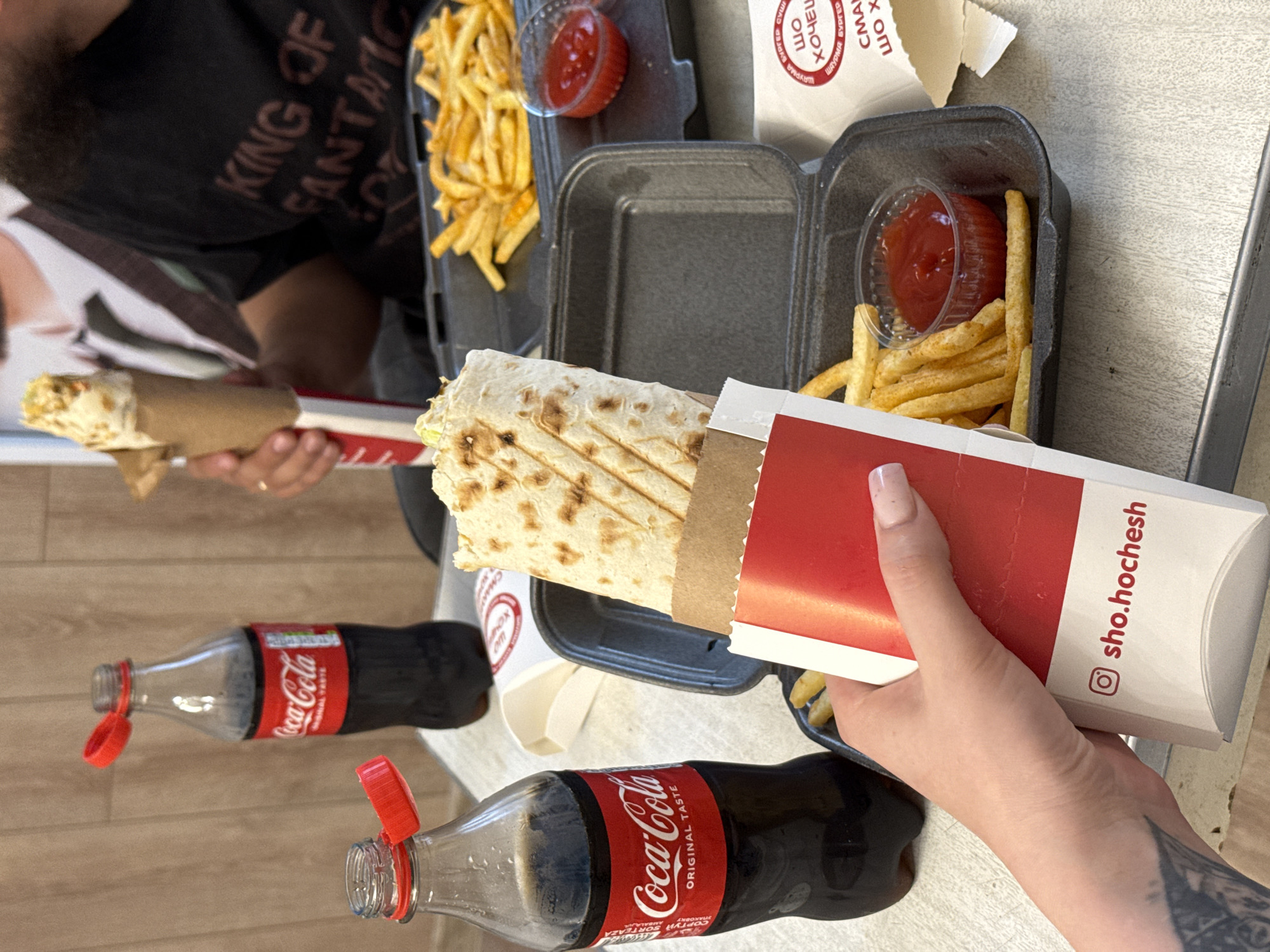A medium shot captures a casual dining scene. In the foreground, a person's hand, adorned with a light pink manicure and a tattoo on the forearm, holds a white paper container with a red band, which contains a shawarma. The shawarma is partially visible, wrapped in a golden-brown flatbread. To the left, a clear plastic bottle of Coca-Cola with a red cap is prominently displayed. Behind it, another identical Coca-Cola bottle stands. In the background, a black takeout container holds a generous portion of golden french fries and a small clear cup of red ketchup. To the right, another person, partially visible, is holding a shawarma. The setting appears to be an indoor establishment, with a light-colored table surface and a wooden-patterned wall in the background. A dark-colored t-shirt with text is also visible on the person in the background. The overall scene suggests a mealtime experience.