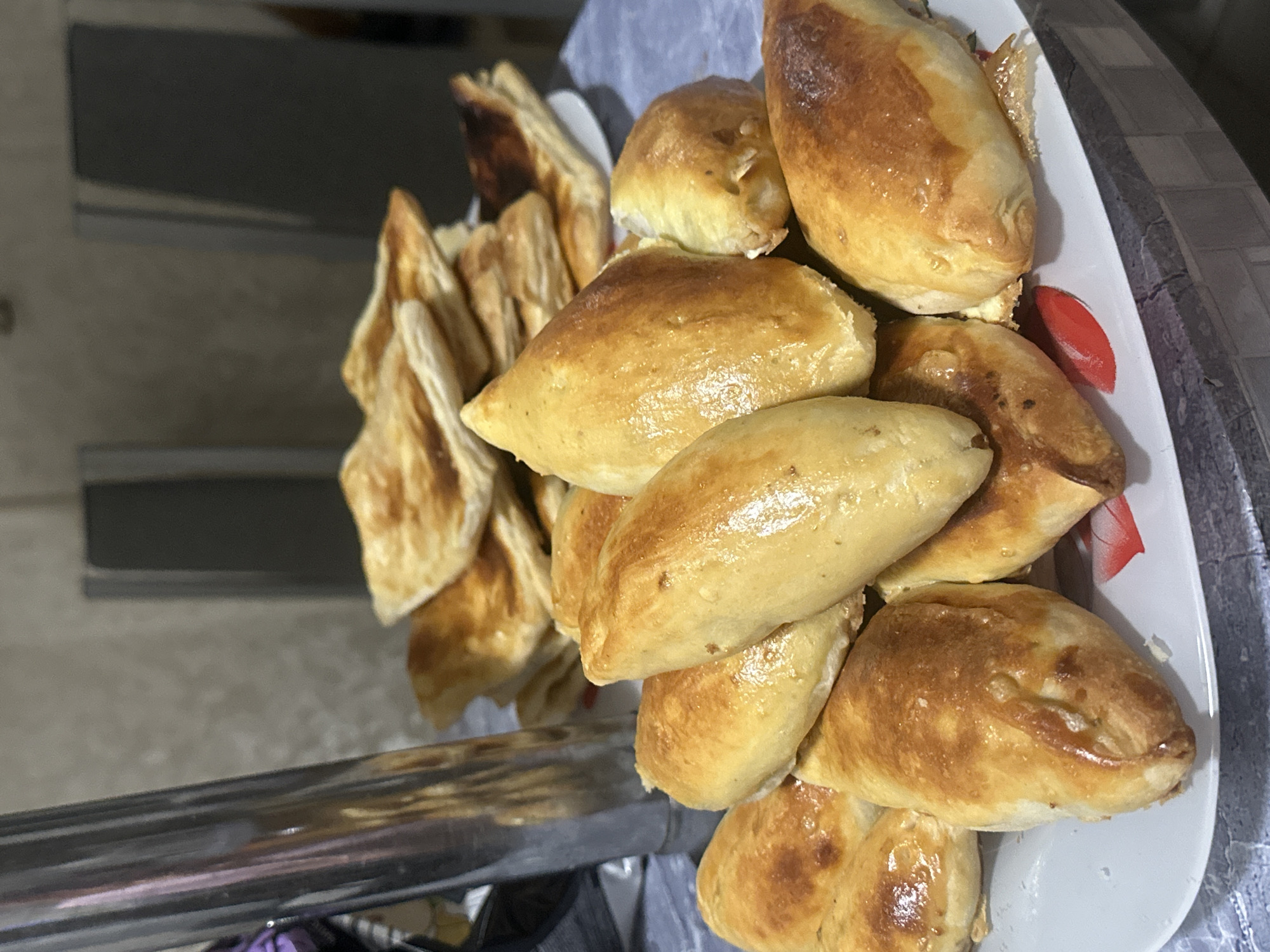 A plate of freshly baked pastries is arranged on a table, likely in a home kitchen, as suggested by the presence of a stainless steel appliance and cabinets in the background, which could be in the city of Perm, Russia. The pastries are golden brown and appear to be filled, with some exhibiting a flaky crust. The plate itself is white with a floral pattern. The lighting is soft, indicating an indoor setting. There are no people or animals visible in the image. The overall impression is one of a home-cooked meal or a snack preparation.