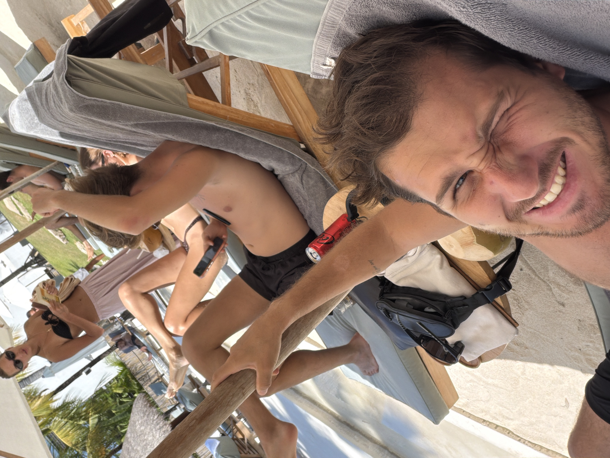 In North Kuta, Indonesia, a group of individuals are relaxing on beach chairs under umbrellas. In the foreground, a man is lying down and looking at the camera, making a funny face. He is wearing black shorts and appears to be on vacation. Behind him, another person is lying on a beach chair, also wearing black shorts. A red can is visible on the chair next to them. Further back, a woman in a black bikini and a light pink skirt is standing and holding something in her hands, possibly a book or magazine. The environment is sandy and sunny, suggesting a tropical beach setting.