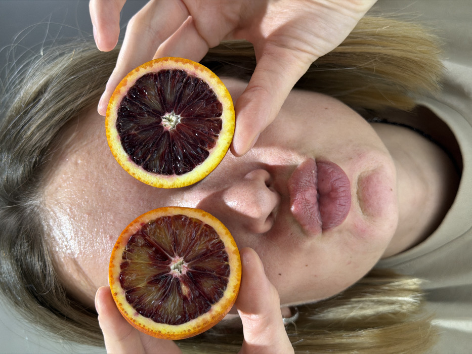 The image depicts a fair-skinned individual, identified as a woman, in an indoor setting in Chaikovsky, Russia. She is shown from the upper chest to just above the forehead, with light brown hair framing her face. Her eyes are obscured by two halves of blood oranges, which she holds in place with her fingers. The oranges display deep red, segmented flesh with a lighter core, surrounded by bright orange-yellow rinds. Her lips are puckered in a "kissing" expression. The background is a plain, light-colored surface, suggesting a minimalist environment.