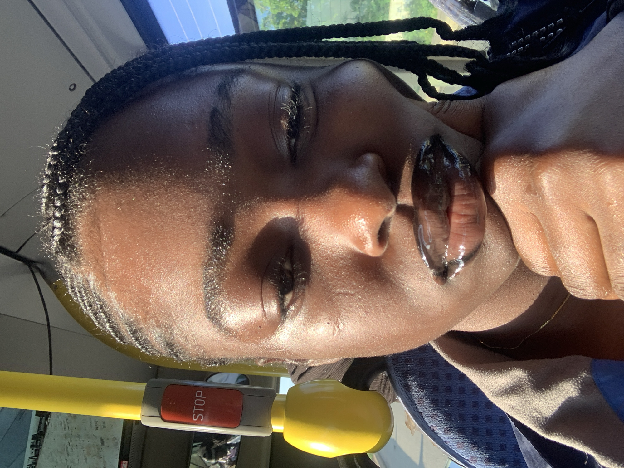 A close-up shot focuses on the face of a Black person with braided hair. They are indoors, likely on a form of public transportation, given the presence of a yellow pole with a red "STOP" button visible on the left side of the frame. The subject has dark skin that appears dewy, with subtle highlights on their forehead and cheeks. Their eyes are partially closed, and their lips are glossy and reflect the light. The background is out of focus, but hints of a window and outdoor greenery suggest a sunny day. The subject's hand is gently resting on their chin, adding a relaxed pose to the composition. The overall lighting is warm and natural, casting soft shadows.