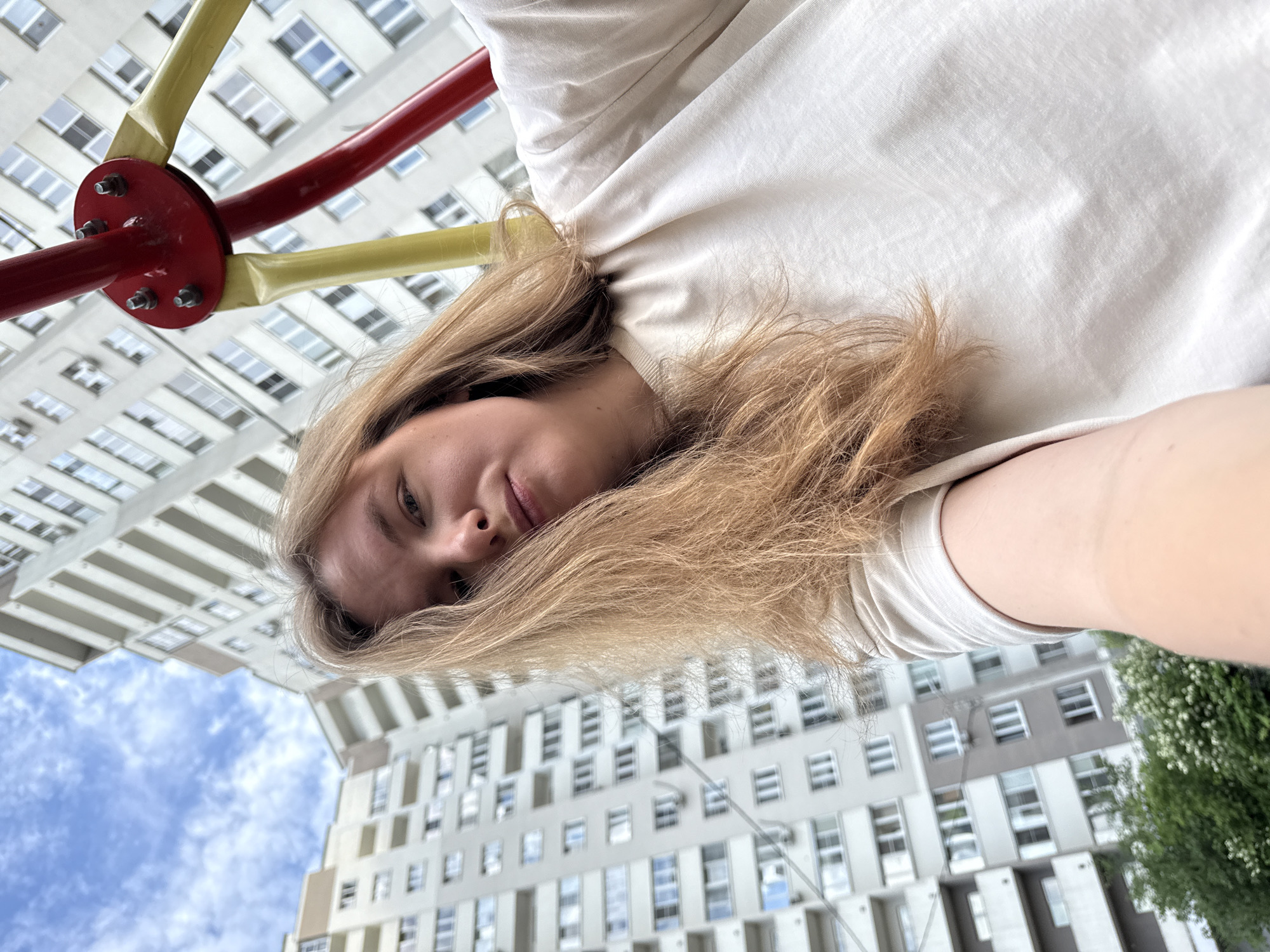 The image is a low-angle selfie taken on a playground with a blurry background of residential buildings and a cloudy sky. A young woman with long, light brown hair is the primary subject, positioned in the foreground. She is wearing a light-colored t-shirt. Part of a red and yellow playground structure is visible in the upper right corner. The residential buildings in the background appear to be tall, multi-story apartment blocks, typical of Soviet-era architecture, and are likely located in the city of Vladimir, Russia, given the context. The lighting suggests it is daytime.