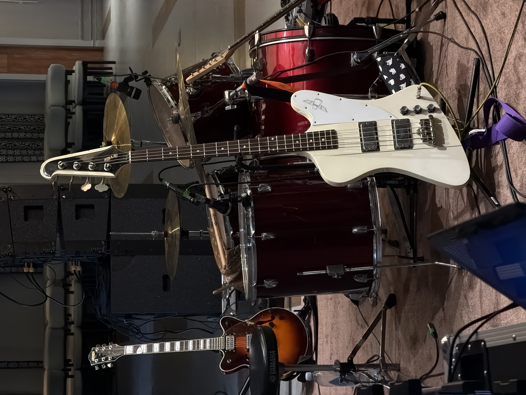 The image displays a collection of musical instruments, primarily a drum set and two guitars, set up in what appears to be a rehearsal space or a small performance venue. A white bass guitar with a distinctive Thunderbird body shape is prominently featured in the foreground, positioned over a maroon drum kit. A second guitar, an electric hollow-body with a sunburst finish, is visible below the bass guitar. The drum set includes various cymbals and drums, with microphones positioned for amplification. Black speaker cabinets are stacked behind the drum kit. Cables and stands are scattered around the equipment, indicating an active setup. The lighting suggests an indoor setting, possibly with artificial light. The overall impression is one of readiness for a musical performance or practice session. There is no visible text in the image, and no people are present. The location context provided is Каджи-Сай 2, Kyrgyzstan.