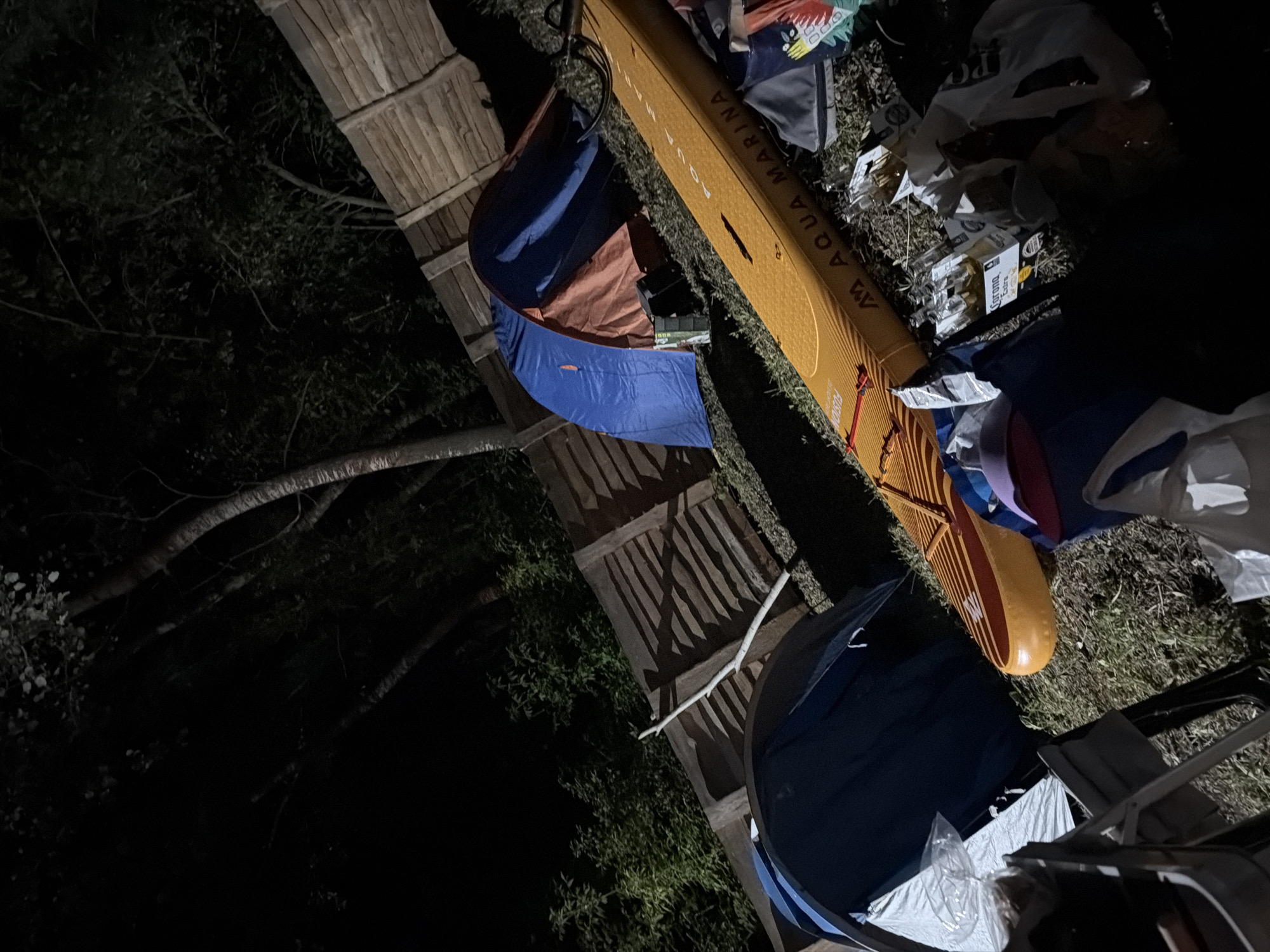 The image depicts an outdoor campsite at night. Two tents are visible, one blue and one blue and orange, set up on grassy ground. A large, orange inflatable paddleboard with the brand name "AQUA MARINA" printed on it lies on the ground next to the tents. Various items are scattered around, including bags, bottles (some appearing to be beer bottles, one labeled "Corona Extra"), and what looks like packaging. A wooden fence is present in the background, with trees and foliage beyond it. The scene suggests a casual camping or outdoor gathering.