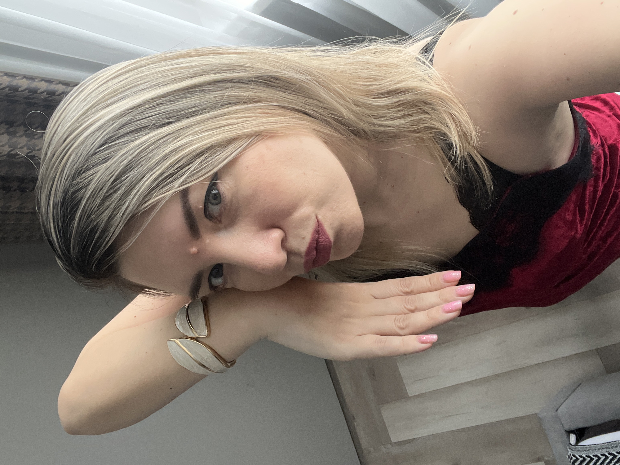 This is a vertical, close-up selfie of a woman with blonde hair and blue eyes. She is wearing a red velvet top with black lace trim. Her left arm is bent, with her hand resting near her face and elbow on a surface. She is wearing a decorative bracelet on her left wrist that appears to be made of gold and white leaf-like shapes. Her fingernails are painted a light pink with glitter. She has on dark lipstick and her expression is subdued. The background shows a blurred interior scene with horizontal blinds and wood-like flooring. The lighting suggests it is daytime. The context provided is Sochi, Russia.