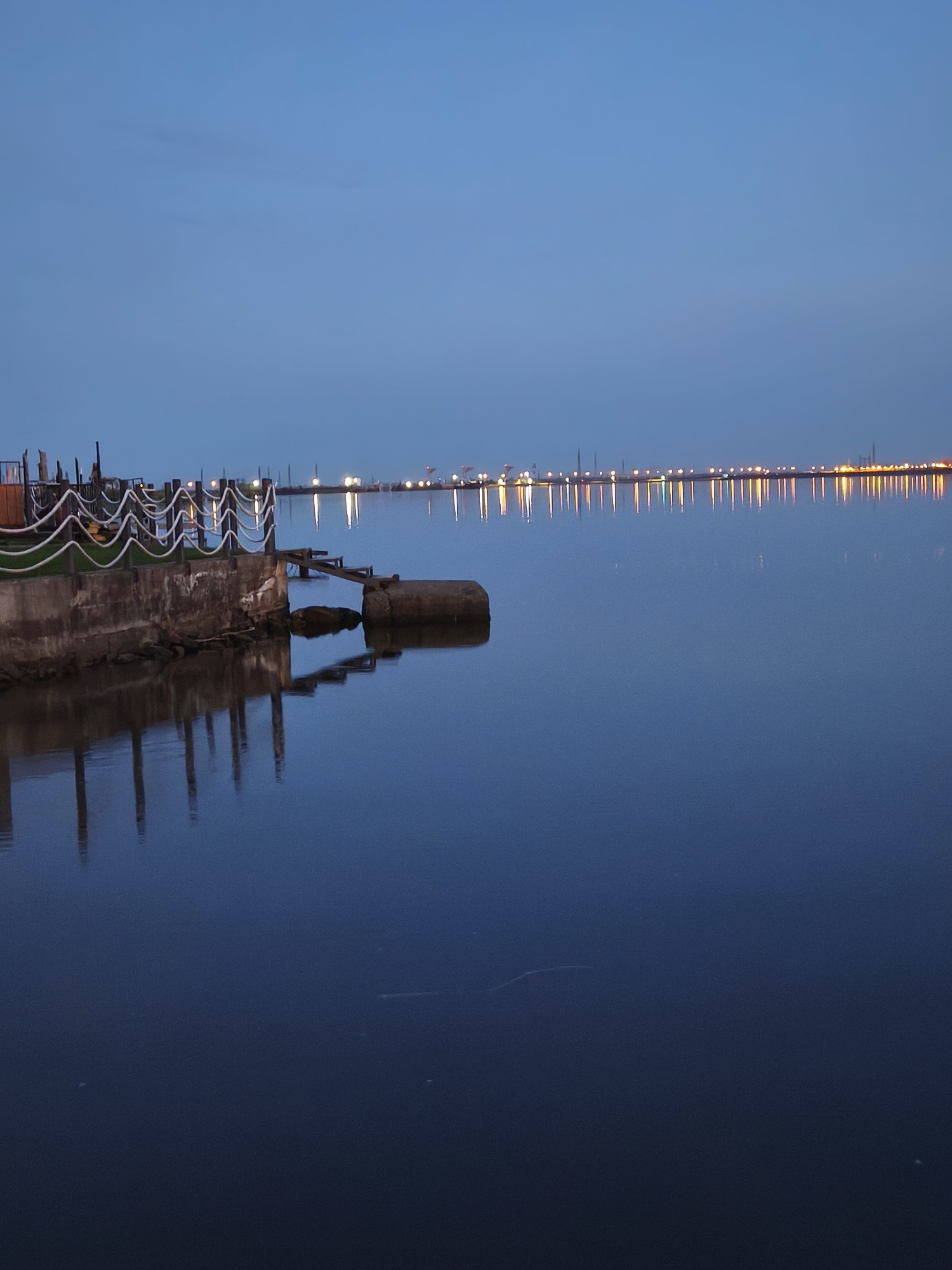 The image depicts a waterfront scene in Chaikovsky, Russia, at dusk or dawn. A concrete pier or embankment with a decorative rope railing extends into a calm body of water. The water reflects the structure and the distant lights along the shoreline. Beyond the pier, a line of lights suggests an urban or industrial area, with various indistinct structures and lights reflected in the water. The sky is a gradient of blue.
