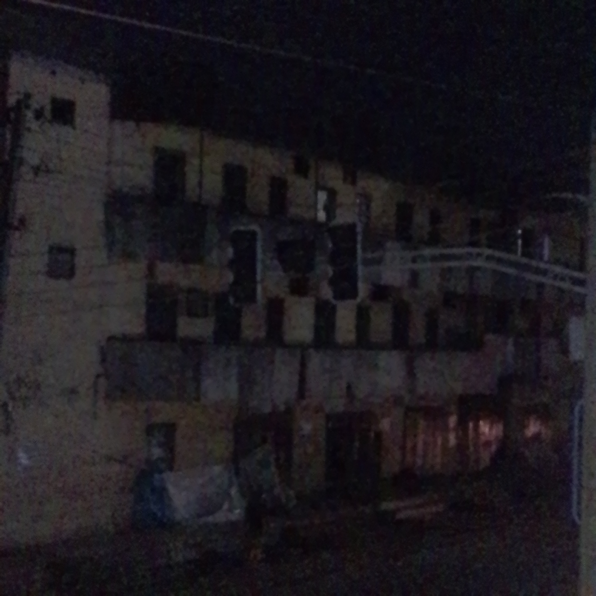 The image depicts a dark, somewhat dilapidated, multi-story building at night. The building appears to be made of concrete or stucco and is a pale yellow or beige color. It has numerous dark rectangular openings that suggest windows or doorways, many of which are empty, revealing the darkness within. Some of these openings have a checkered pattern of light and dark, possibly indicating internal structures or shading.
The building has a layered appearance, with balconies or recessed sections on the upper floors, supported by visible beams or columns. There are some piles of debris or materials visible in the foreground at the base of the building, including what looks like a blue tarp or covering. The overall impression is one of neglect or disrepair.
The scene is illuminated by a low-light setting, indicating it is nighttime, and there are no clear indications of specific weather conditions. The location cues from the provided context place this scene in Maiburiji, Nigeria. There are no visible people or animals in the image, and no text is discernible. The atmosphere conveyed is somber and perhaps slightly eerie due to the darkness and the state of the building. A curved structure, possibly a ramp or walkway, is visible on the right side of the building in the background, adding another architectural element to the scene.