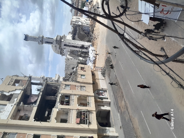 An aerial perspective depicts a heavily damaged urban street in Khan Yunis, Palestinian Territory, under a partly cloudy sky. On the left, a multi-story building exhibits severe structural damage, with upper floors collapsed, exposed interiors, and lower-level balconies containing hanging laundry. Adjacent to this, in the background-right, a mosque with a domed roof and a tall minaret stands partially destroyed, surrounded by extensive rubble. The paved street running through the center features faded white lane markings. Scattered across the street and sidewalks are approximately a dozen individuals, predominantly males, engaged in walking or standing. In the immediate foreground, two individuals walk on the street. Towards the right mid-ground, a small group of individuals is seated amidst a sandy, rubble-strewn area. A white banner with red Arabic script is visible in the far right foreground, partially obscured by a network of utility wires and metal structures. The entire scene is characterized by widespread debris and structural wreckage.