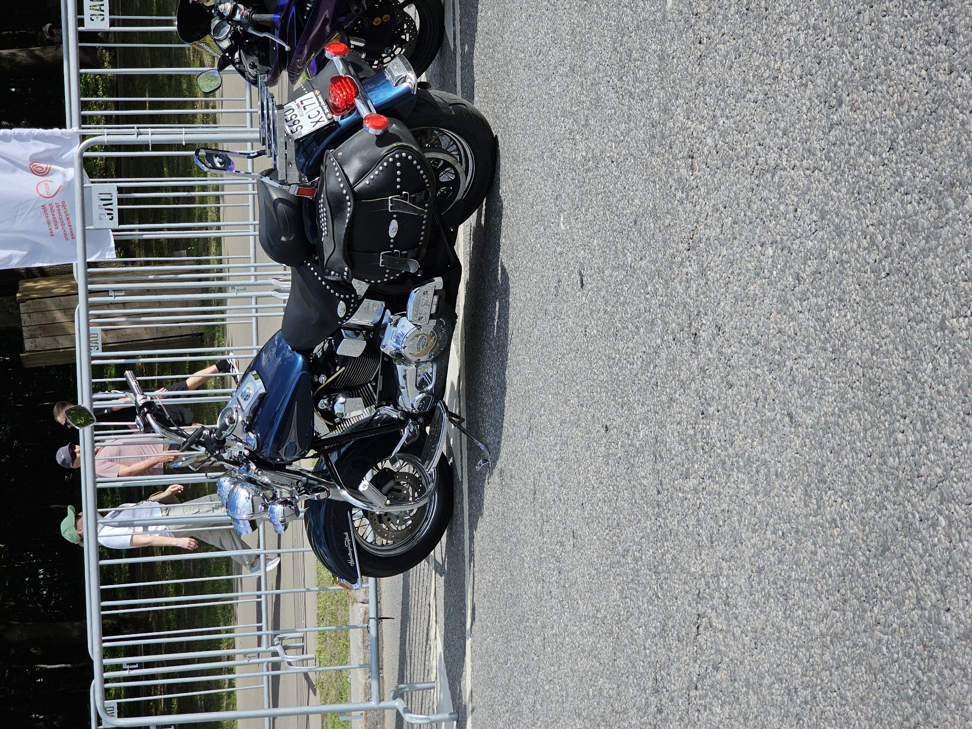 The image shows two motorcycles parked on an asphalt surface, situated next to a metal barrier. The motorcycle closer to the camera is a blue and chrome model, equipped with studded leather saddlebags and a license plate displaying "5650 XC77 RUS". The second motorcycle is a purple color. Behind the barrier, three individuals are partially visible. The location is a street in the city of Unknown, Unknown.
