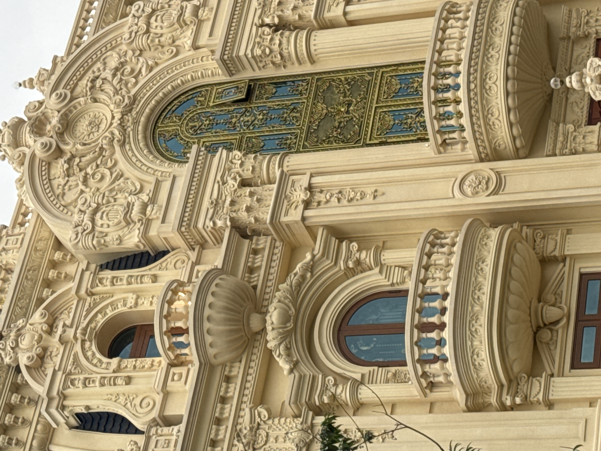 The image depicts a close-up view of the ornate facade of a building in Da Lat, Vietnam. The architecture is characterized by elaborate decorative carvings, predominantly in a creamy yellow hue. Prominent features include arched windows with dark frames, balconies with detailed balustrades, and intricate sculptural elements adorning the structure. One large, tall window with a blue and gold decorative pattern is visible on the right side of the image. There are no people or active interactions visible. The environment appears to be an exterior architectural setting.