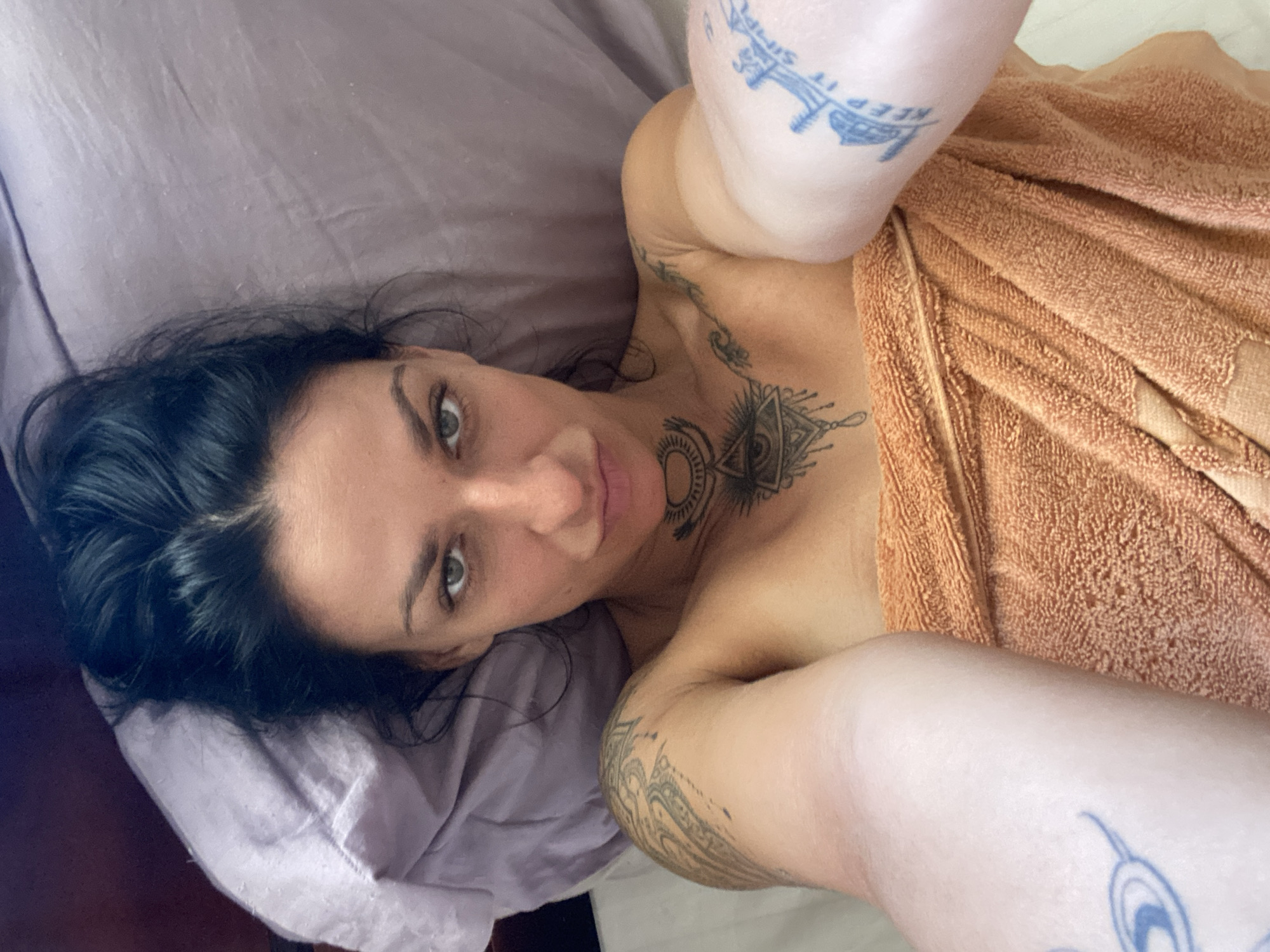 A person with dark hair and light eyes is lying on a pillow. The person has several tattoos on their arms, chest and neck. An orange towel is draped over the person's body. The location is not discernible from the image.