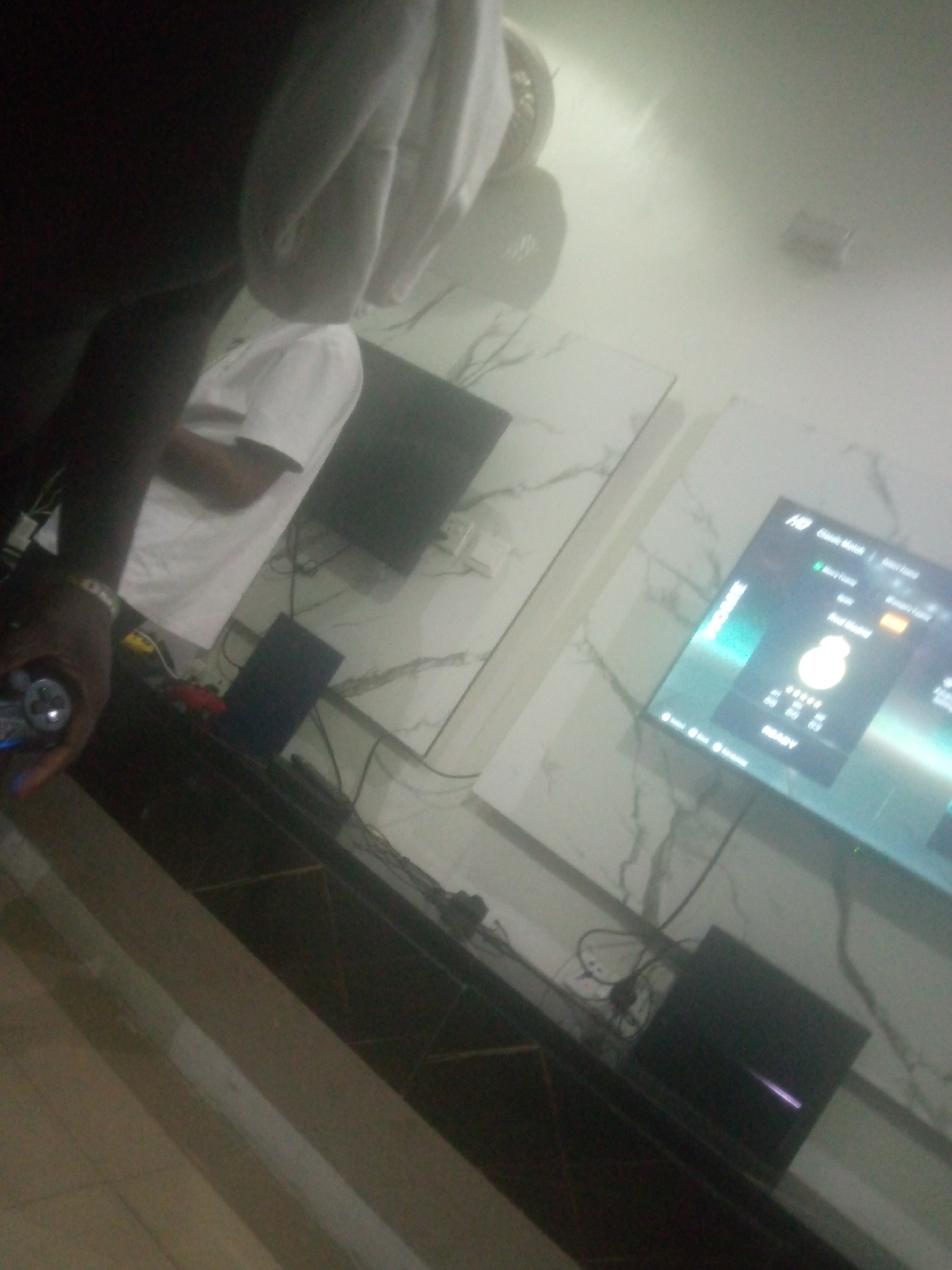 The image depicts an indoor scene, likely a living room or entertainment area, with a television screen displaying a video game interface. The television is mounted on a wall with a marble-patterned finish. In the foreground, a person is partially visible, holding a video game controller, suggesting active gameplay. Other electronic devices, possibly gaming consoles and accessories, are situated on a dark wooden entertainment unit beneath the television. The overall ambiance appears casual and focused on entertainment. The image quality is low, making it difficult to discern fine details of the people or objects. No definitive landmarks or elements within the image can be used to ascertain the specific location within Jalingo, Nigeria.