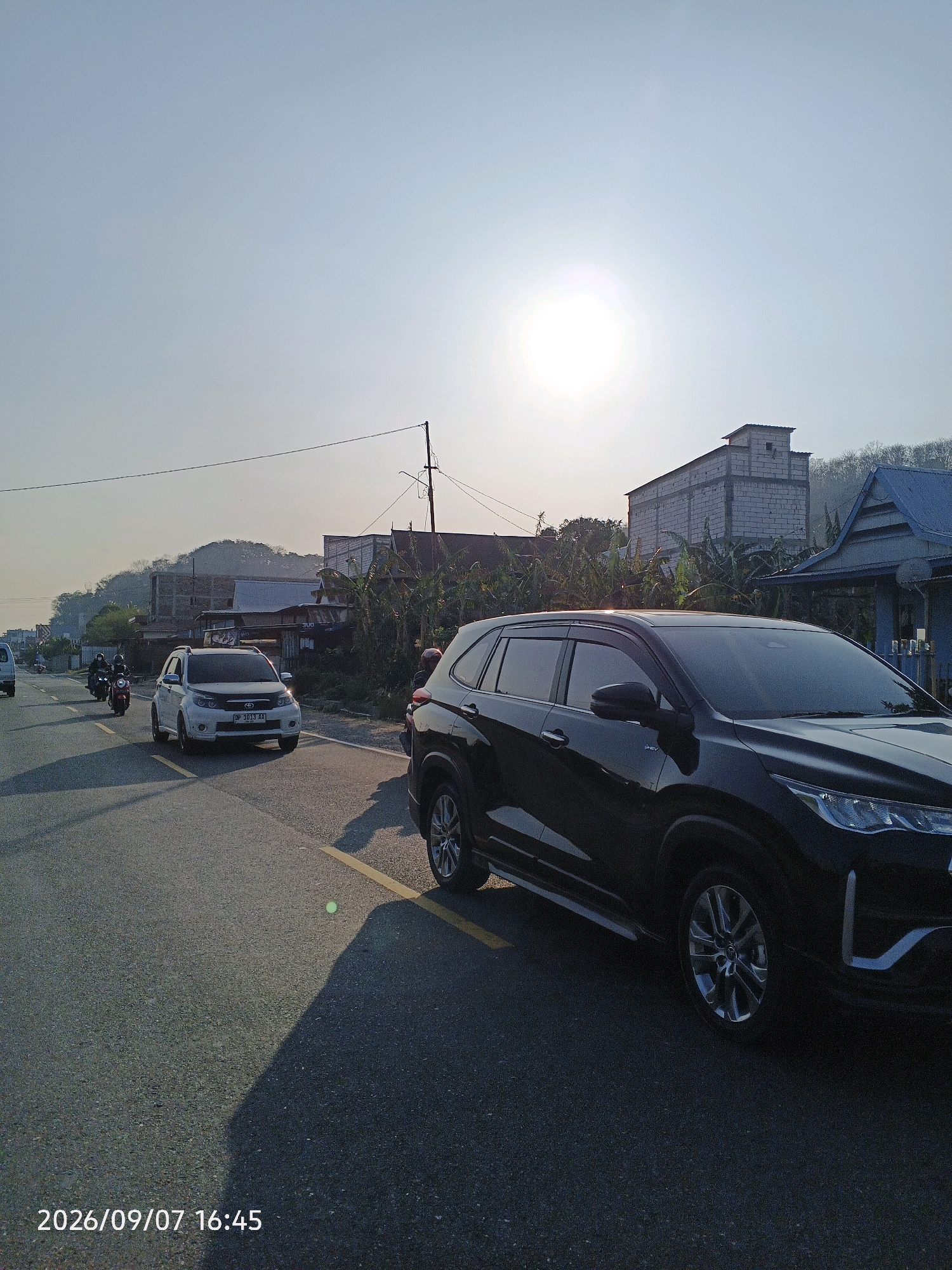 The image captures a street scene in Lainungan, Indonesia, on a sunny day. A black SUV is parked on the right side of the road, facing away from the viewer. The sun is high in the sky, casting a bright light and long shadows. In the background, a white car is driving away from the camera, followed by two motorcycles. Buildings and trees line the road, with a hill visible in the distance. The timestamp "2026/09/07 16:45" is visible in the bottom left corner.