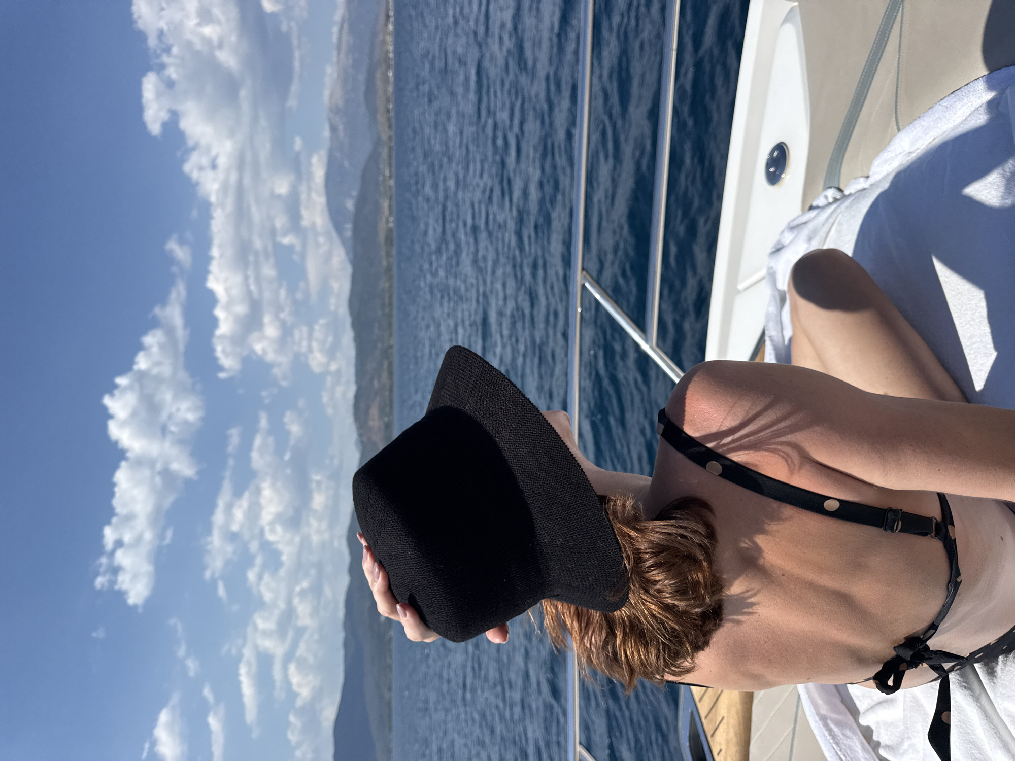 A person is shown from the back, wearing a black hat and a black bikini. They are sitting on a white towel on a boat, with their legs extended. The boat appears to be on the sea, with blue water stretching out to the horizon. In the distance, there are mountains and a clear blue sky with fluffy white clouds. The sun is shining, casting shadows on the boat and the person. The overall scene suggests a leisurely day spent on a boat, enjoying the sea and the sunshine. The person's posture, with their head down and hand on the hat, could imply relaxation or seeking shade from the sun. The clear sky and bright sunlight indicate it is daytime, likely a warm and pleasant day. The presence of mountains in the distance suggests a coastal location.