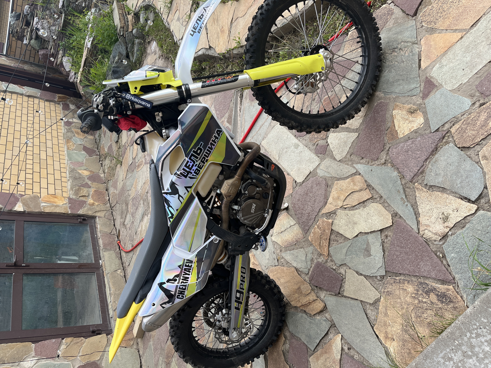 A Husqvarna dirt bike, possibly an enduro model, is prominently featured in this outdoor, vertically oriented image. The motorcycle, with its distinctive yellow front fender and fork guards, is parked or leaning against a rustic stone wall. The bike's bodywork features a holographic or iridescent graphic design with black and white patterns.
Visible text on the bike includes "Husqvarna" on the handlebar clamp, "HTW SUSPENSION" on the fork tube, and "H9 PRO" on the lower fork. Several decals feature Cyrillic text: "ЦЕЛЬ-ВЕРШИНА" (Tsel-Vershina, meaning "Goal-Peak") is seen on the tank shroud and front fender, and "CHERNYAEV" is present on the side fairing, likely a rider's name. An "SN" logo is visible on the engine cover. The bike has knobby tires, suitable for off-road riding, and a black textured seat. A small red cloth or bag is tucked near the handlebars.
The background consists of a multi-colored stone facade that extends from the ground up, forming part of a building wall. A brown-framed window is visible on the building to the left. Above the stone wall, lush green foliage and rocks indicate a garden or natural landscape. String lights with small, clear bulbs are strung across the top left of the image. The ground in the foreground is paved with irregularly shaped flagstones in various earthy tones. The scene appears to be set during daytime with clear weather, likely in Ореховка, Russia.