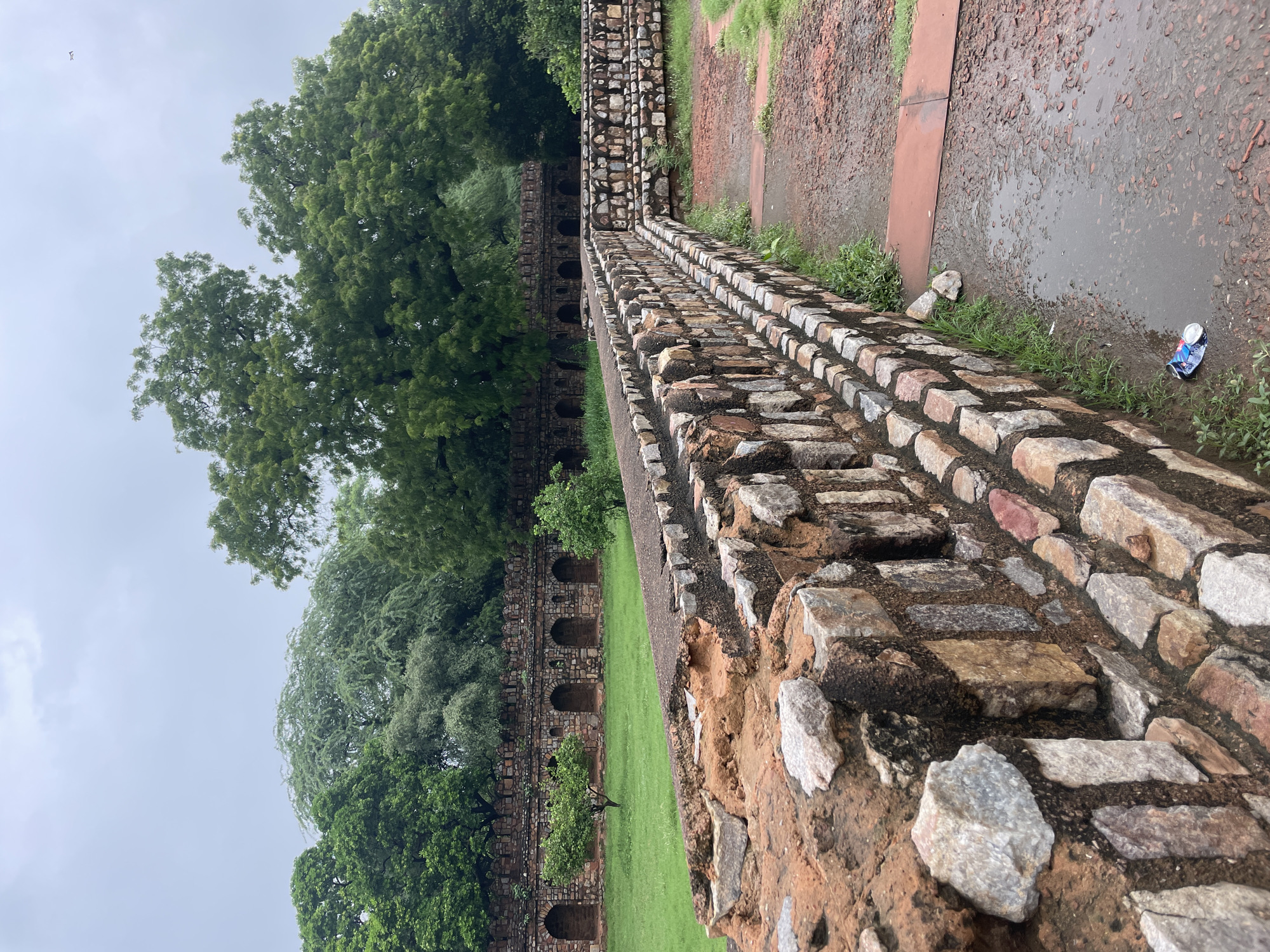 This is a photograph of the tomb of Safdarjung in Delhi, India. The image captures a section of the garden and the surrounding walls. In the foreground, a rough stone wall with a weathered, reddish-brown mortar is visible. This wall extends to the right, creating a pathway of gravelly, reddish-brown earth. A puddle of water is present on the ground, with a crushed aluminum can lying next to it.
In the middle ground, another stone wall runs horizontally across the frame. This wall is constructed from smaller, more regularly shaped stones and features a series of arched alcoves along its length. Lush green grass covers the ground in front of this wall. Two small trees are planted in the grassy area.
The background is dominated by a dense canopy of green trees, suggesting a humid and possibly rainy day, given the presence of the puddle and overcast sky. The overall impression is one of a historical site within a verdant parkland.