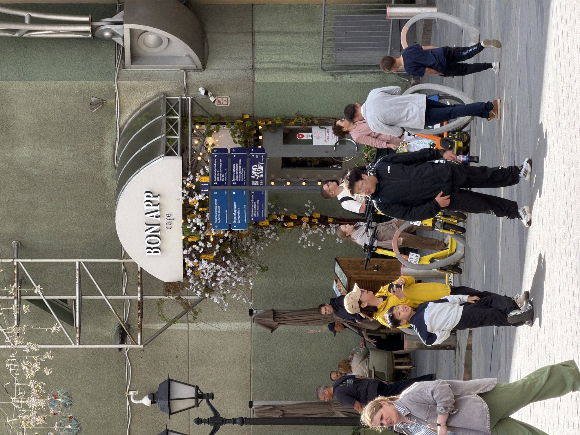 In this outdoor scene, a group of people are gathered in front of a cafe named "BON APP cafe." The cafe has a white awning with the name prominently displayed and a glass-paneled entrance. Yellow flowers and greenery are arranged around the entrance. Several signs with blue backgrounds and white text are posted to the right of the entrance, indicating directions to various locations like the Polytechnic Museum, Zaryadye Park, and a post office.
In the foreground, a young boy in a white and blue tracksuit stands with his hands on his hips, facing forward. To his left, a woman in a yellow hooded jacket holds a coffee cup. Further left, a woman in a grey jacket and green pants walks away from the camera.
In the midground, several people are seated at tables outside the cafe, partially obscured by umbrellas. Others are standing and interacting. Two yellow electric scooters are parked to the right of the boy.
In the background, the building facade is a muted green color. A street lamp with multiple bulbs is visible on the left side of the image. The ground is paved with light-colored tiles. Shadows from the people and objects are cast on the ground.
