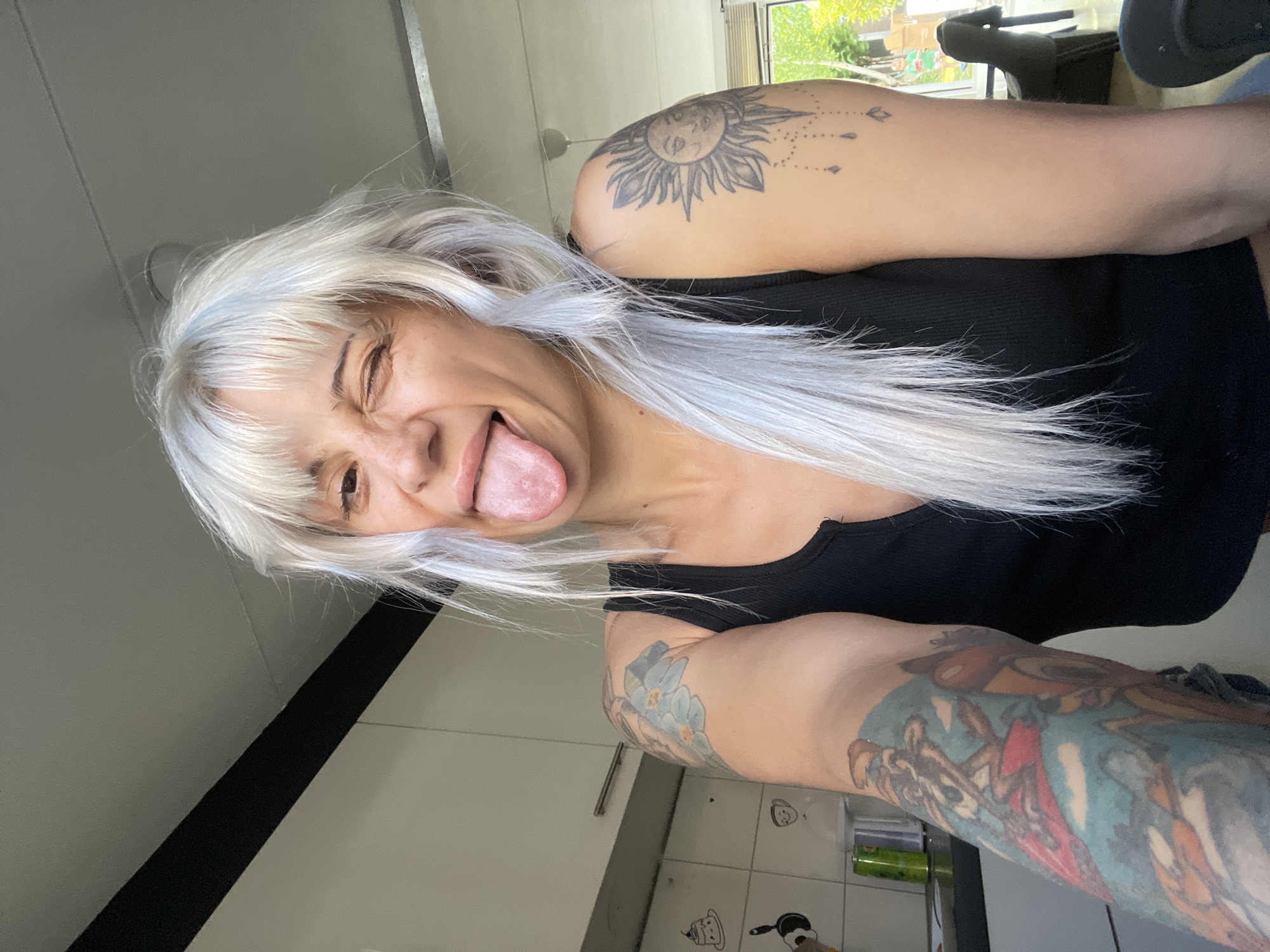 A woman with white hair and tattoos on both arms sticks out her tongue, winking. She wears a black tank top. The background reveals a kitchen with white cabinets and tile. A window is visible, showing greenery and a building in the city of Tilburg, Netherlands. A black office chair is partially visible through the window.
