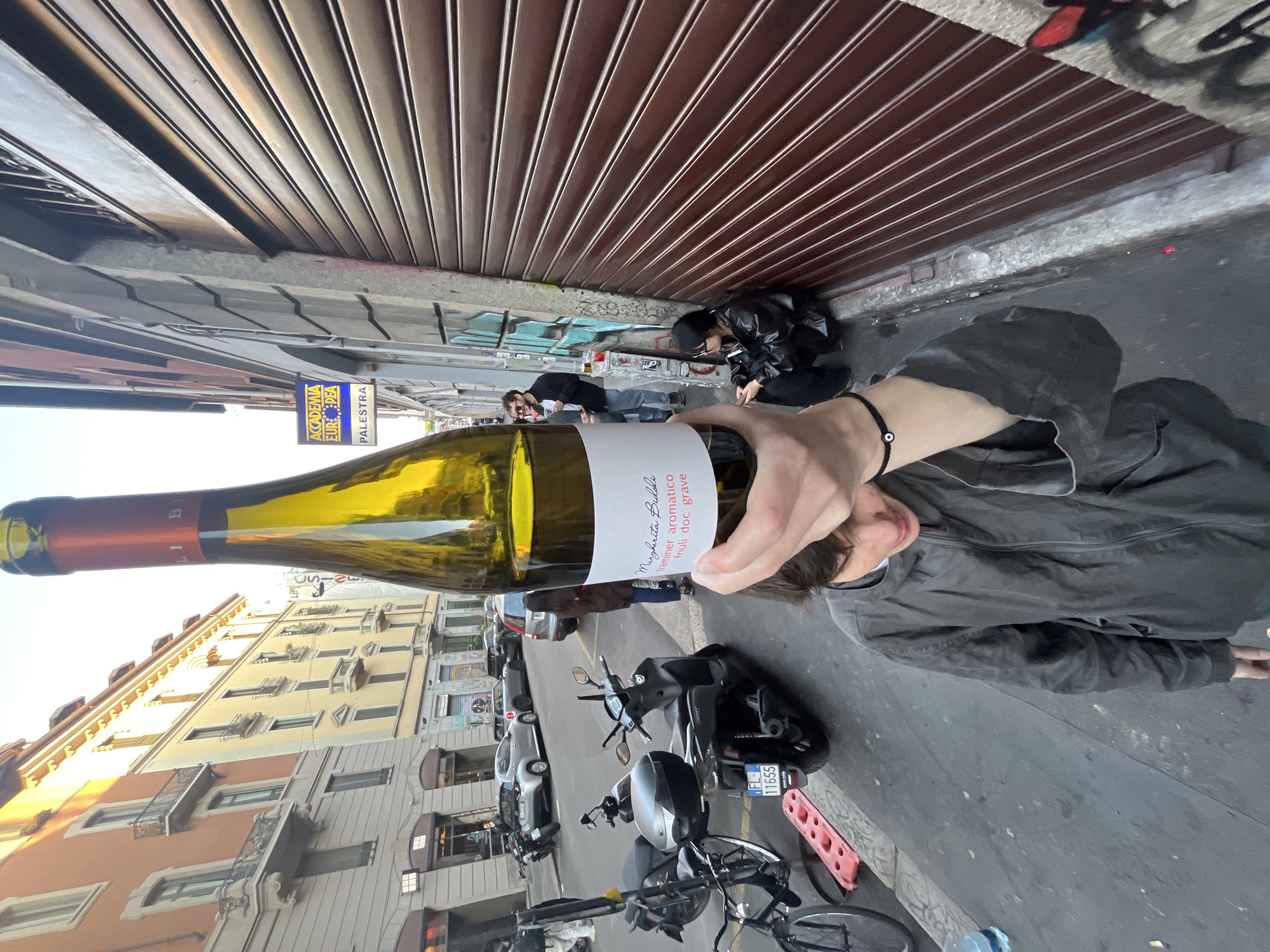 The image, rotated ninety degrees counter-clockwise, captures a casual street scene in an urban environment during what appears to be a bright, clear day. In the immediate foreground, a person's hand holds a bottle of white wine, partially obscuring their face. A black bracelet with an evil eye charm is visible on their wrist. The wine label clearly reads "Margherita Bidoli traminer aromatico friuli doc grave."
In the midground, two individuals are present near a closed brown metal storefront shutter. One person, dressed in a black jacket, is crouched or sitting on the ground. Another person stands further back against a wall with graffiti. Further down the street, another person is seen near a blue and yellow sign.
The street is lined with parked vehicles, including several motorcycles and scooters, one with a license plate reading "FL 11655," and cars. Multi-story buildings with balconies line the street, characteristic of European architecture. A red plastic barrier and a discarded water bottle are on the sidewalk. Visible text on a sign includes "ACCADEMIA EUROPEA PALESTRA" (European Academy Gym) and "LPM." The scene suggests people are gathered or passing through a bustling street, possibly socializing.