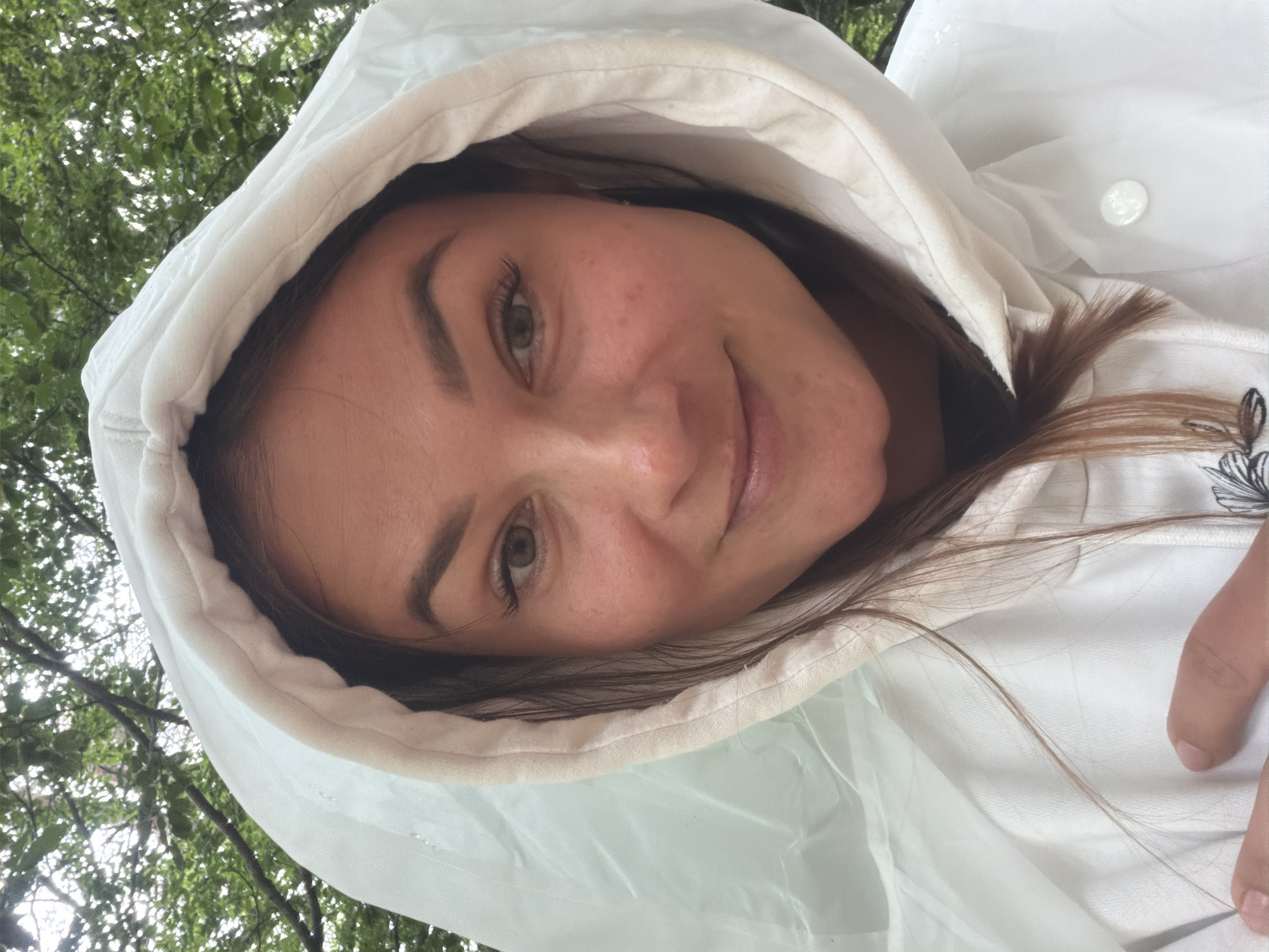 A young woman is captured in a close-up, tilted selfie, wearing a white hooded raincoat. She has brown hair, prominent eyebrows, and green or hazel eyes, with freckles visible across her cheeks and nose. Her expression is a subtle, closed-mouth smile, conveying a pleasant or content mood. The hood frames her face, with a softer, off-white lining visible against her skin and a more plastic-like, translucent outer material for the main body of the raincoat. A single white snap or button is visible on the raincoat near her shoulder.
The background consists of blurred green foliage, indicative of trees or dense vegetation, suggesting an outdoor setting. The lighting appears soft and diffused, characteristic of an overcast day or dappled light filtering through leaves. Strands of her brown hair are visible outside the hood. The overall scene suggests she is outdoors, possibly protecting herself from the elements in Mali Bostur, Montenegro. No text is visible in the image.