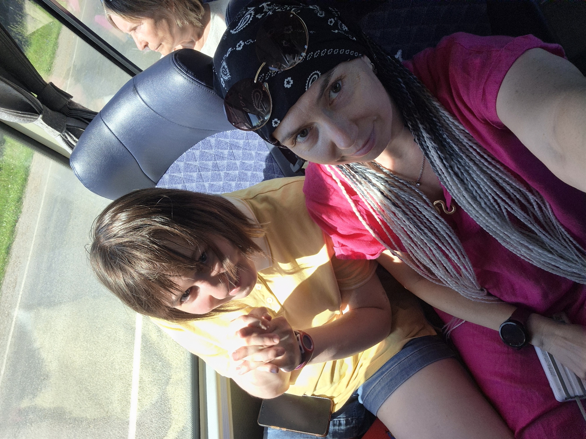 Two women are seated on a bus. The woman in the foreground has braids and is wearing a pink shirt and bandana. The woman in the background is wearing a yellow shirt and denim shorts. The bus appears to be moving, with a glimpse of green grass and pavement visible through the window. The lighting suggests it is daytime.