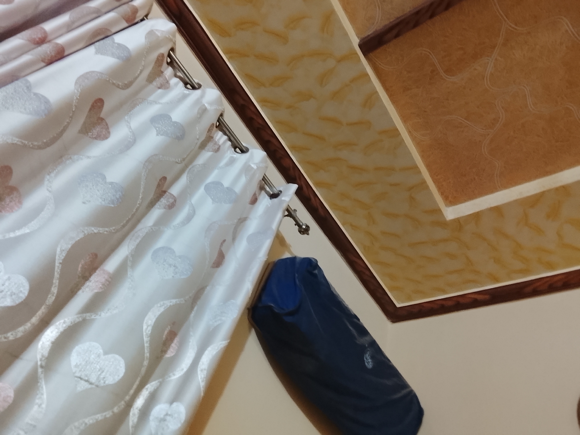 The image shows a portion of a room, likely in Multan, Pakistan, given the prompt's specification. White curtains with heart patterns hang from a metal rod with hooks. Beside the curtains is a folded blue garment. The ceiling has decorative panels in brown and beige tones with a dark wooden trim. The walls are painted in a plain, light color. There are no people visible in the frame.