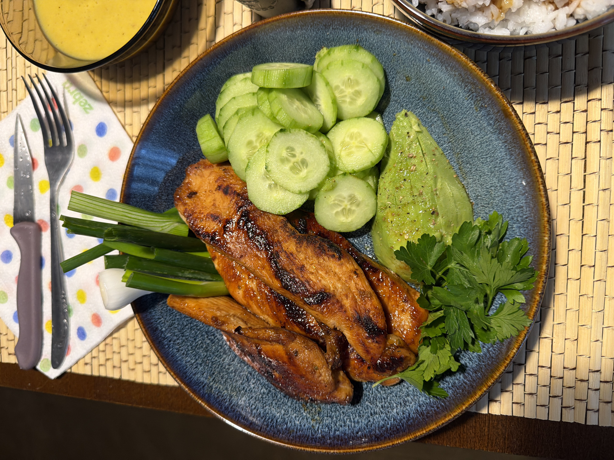 The image displays a fresh and inviting meal, presented on a round, textured blue ceramic plate. The main components of the dish include several pieces of grilled protein, possibly chicken or fish, exhibiting a glossy, caramelized coating and distinct char marks. These are artfully arranged alongside neatly sliced cucumbers, a substantial wedge of avocado seasoned with ground black pepper, and several stalks of vibrant green onions. A small bunch of fresh parsley garnishes the plate, adding a touch of green.
The plate rests on a light-colored, woven placemat, likely made of bamboo or straw. To the upper left, a clear glass bowl contains a creamy, yellow sauce or dressing. Partially visible in the upper right is a metallic bowl holding white rice, possibly with a light seasoning or sauce. A white paper napkin with cheerful multicolored polka dots (red, blue, green, orange, yellow) is positioned to the left of the main plate, holding a silver fork and a knife with a grey handle. The word "Zebra" is visible in reversed text on the napkin.
Strong, warm light, possibly from a window or a direct light source, illuminates the scene from the right, creating defined shadows and highlighting the textures of the food and the placemat. This suggests a daytime setting, likely indoors. The overall presentation indicates a well-prepared and healthy meal, ready for consumption in a casual setting, potentially in Varna, Bulgaria. No individuals are visible in the image.