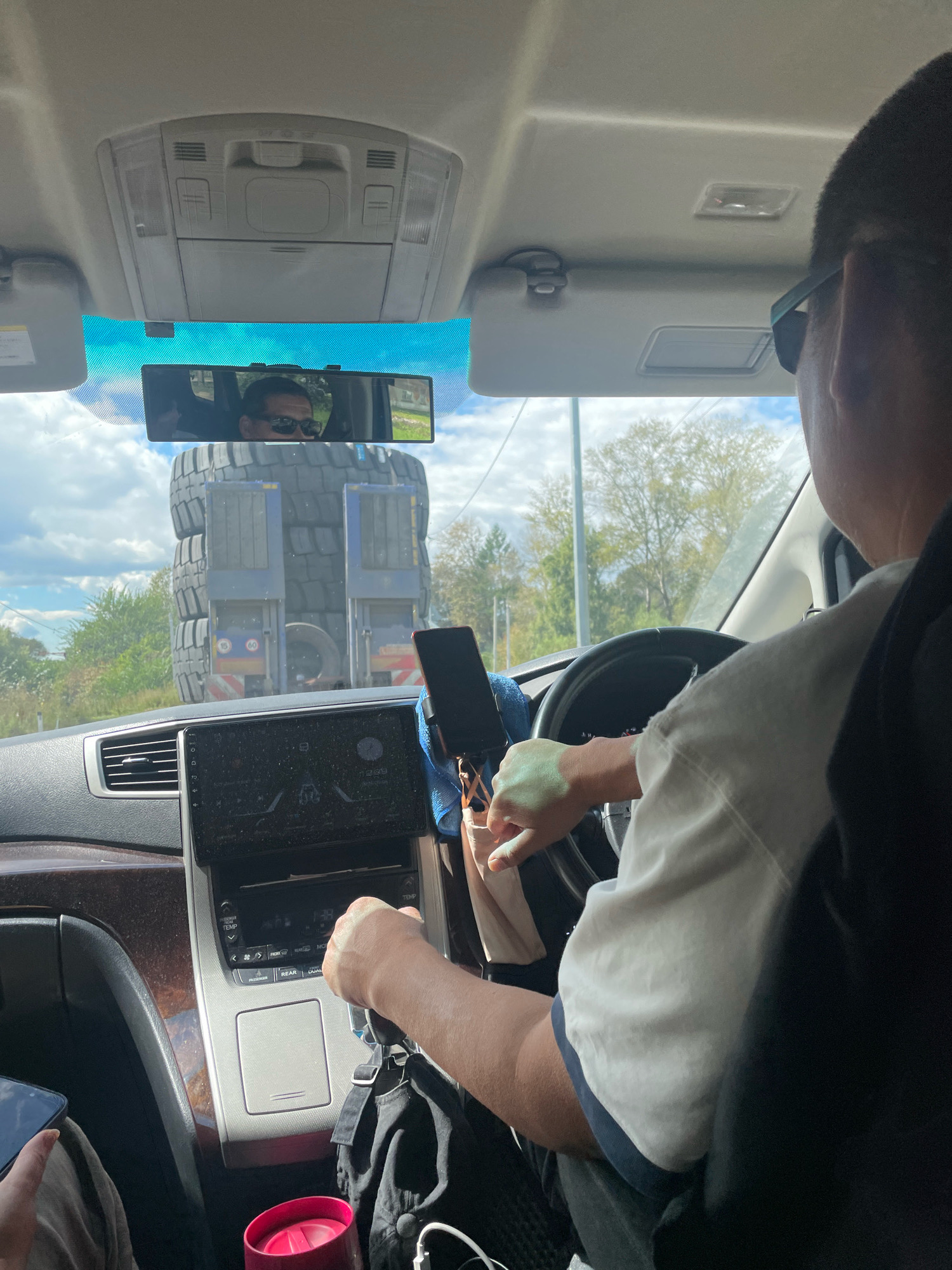 The image shows the interior of a car, with the driver's perspective looking forward through the windshield. In the foreground, the steering wheel and dashboard are visible. A hand is on the gear shift. The driver, seen from the side, is wearing a white and blue shirt. A phone is mounted on the dashboard. In the rearview mirror, the driver's reflection is visible, wearing sunglasses. Outside the car, a large truck carrying massive tires is visible on a road. The scene appears to be in an outdoor, possibly rural setting with trees and a cloudy sky. The location is identified as Budukan, Russia.