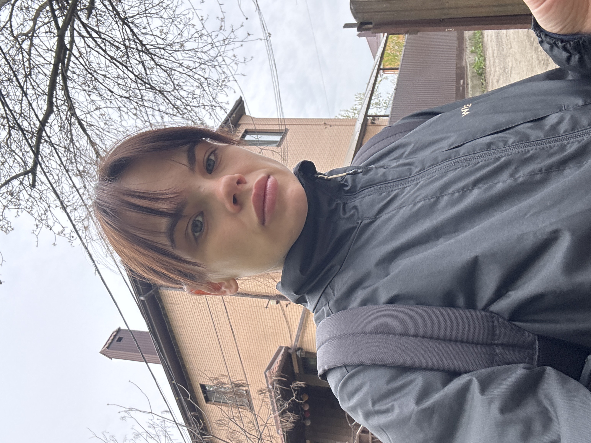 A close-up, low-angle selfie shows a person with brown hair and bangs wearing a dark gray jacket and a gray backpack. The person's face is centered in the frame, revealing clear skin and light-colored eyes.
Behind the person, a two-story brick building is visible, and bare tree branches reach towards the sky. Utility poles and wires crisscross above the building. The sky is overcast. A weathered wooden fence runs vertically on the right side of the frame.
While Rostov-on-Don, Russia, is specified as the location, no visual cues in the image confirm this information. The setting appears to be a typical residential area.