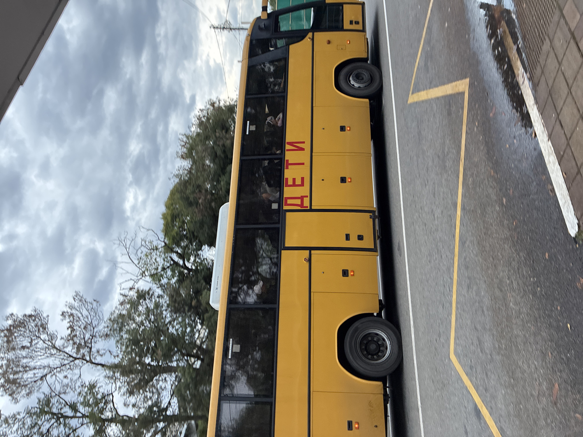 A yellow school bus is parked on a wet asphalt road with yellow markings. The bus has several windows, some of which show the silhouettes of children inside. The word "ДЕТИ" (children) is written in red letters on the side of the bus. Trees and a cloudy sky are visible in the background. The scene appears to be in Sochi, Russia.