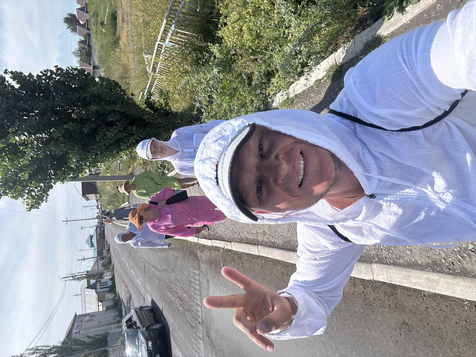A selfie shows a man in a white hooded jacket with a black backpack strap visible, holding up his left hand in a peace sign. He is on a paved road with a sidewalk. Behind him, four people are walking away from the camera. The first person is a woman in a pink long-sleeved shirt and an orange hat. Next to her is another woman in a green long-sleeved shirt and a tan hat. Further back, a person in a white hooded shirt and a white cap is walking. The road is lined with trees and buildings, including some houses with red roofs in the background. A dark-colored car is parked on the left side of the road. The environment appears to be rural or suburban, consistent with a location in Zharbulak, Kazakhstan. The sky is partly cloudy.