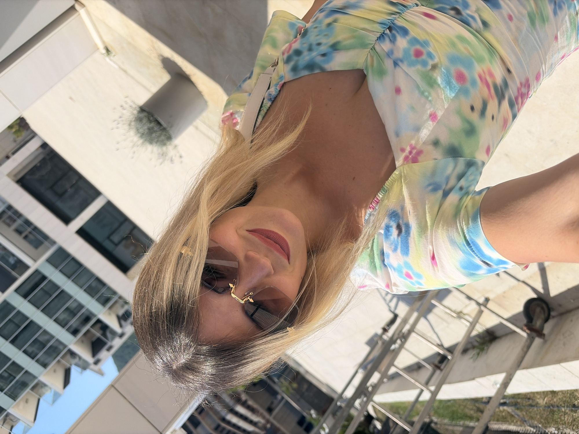 A young woman with blonde hair and sunglasses is taking a selfie outdoors in Budva, Montenegro. She is wearing a tie-dye dress with blue, green, pink, and yellow colors. In the background, a modern building with many windows and a blue sky are visible. The lighting suggests it is daytime, likely sunny.