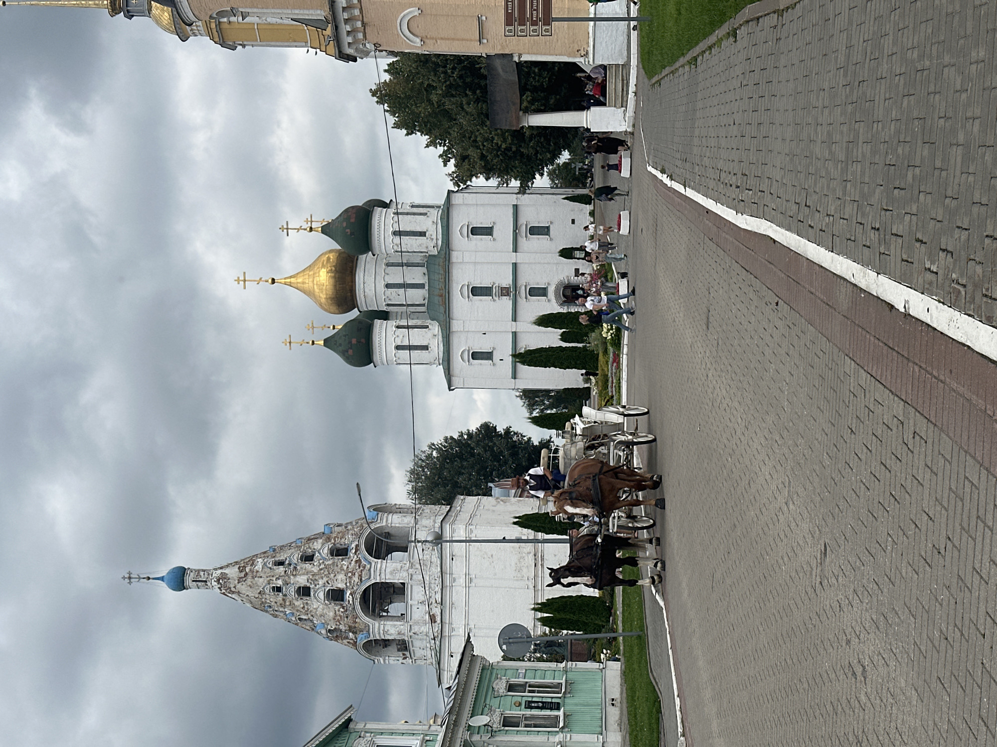 The image depicts a view of the Kolomna Kremlin in Kolomna, Russia, under an overcast sky. In the foreground, a cobblestone road leads towards a complex of churches. A horse-drawn carriage, with two horses and a driver wearing a hat and vest, is moving along the road. To the left, a light green building with white trim is visible. To the right, a large white church with multiple golden domes and green accents stands prominently. Behind it, a bell tower with a blue dome and ornate detailing is partially obscured by trees. Further to the right, a section of another building with a tall golden spire can be seen. Several people are walking around the church complex, some standing near the entrance and others in the plaza. The overall scene suggests a tourist attraction or a place of historical and religious significance.