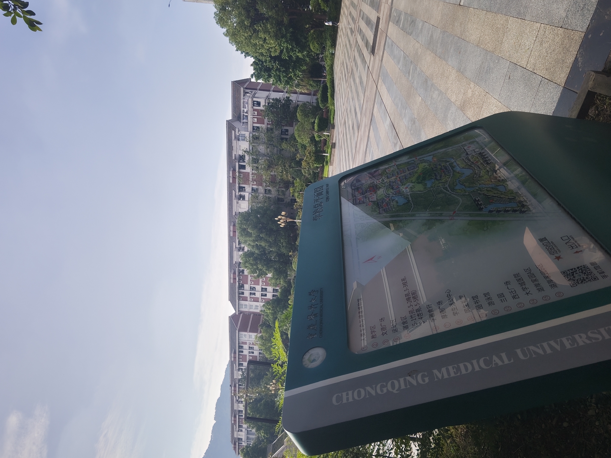 A green information kiosk with a map is situated in front of several multi-story buildings and trees under a clear sky. The kiosk displays Chinese text, including "CHONGQING MEDICAL UNIVERSITY," a campus map, and a list of locations. The ground in front of the kiosk is paved with rectangular stones in a striped pattern. A person in a white shirt and dark shorts is walking on the right side of the paved area, away from the camera. The buildings appear to be part of a university campus, with windows and balconies visible. The overall scene suggests a bright, sunny day on a campus in the city of Chongqing High-tech Zone, China.