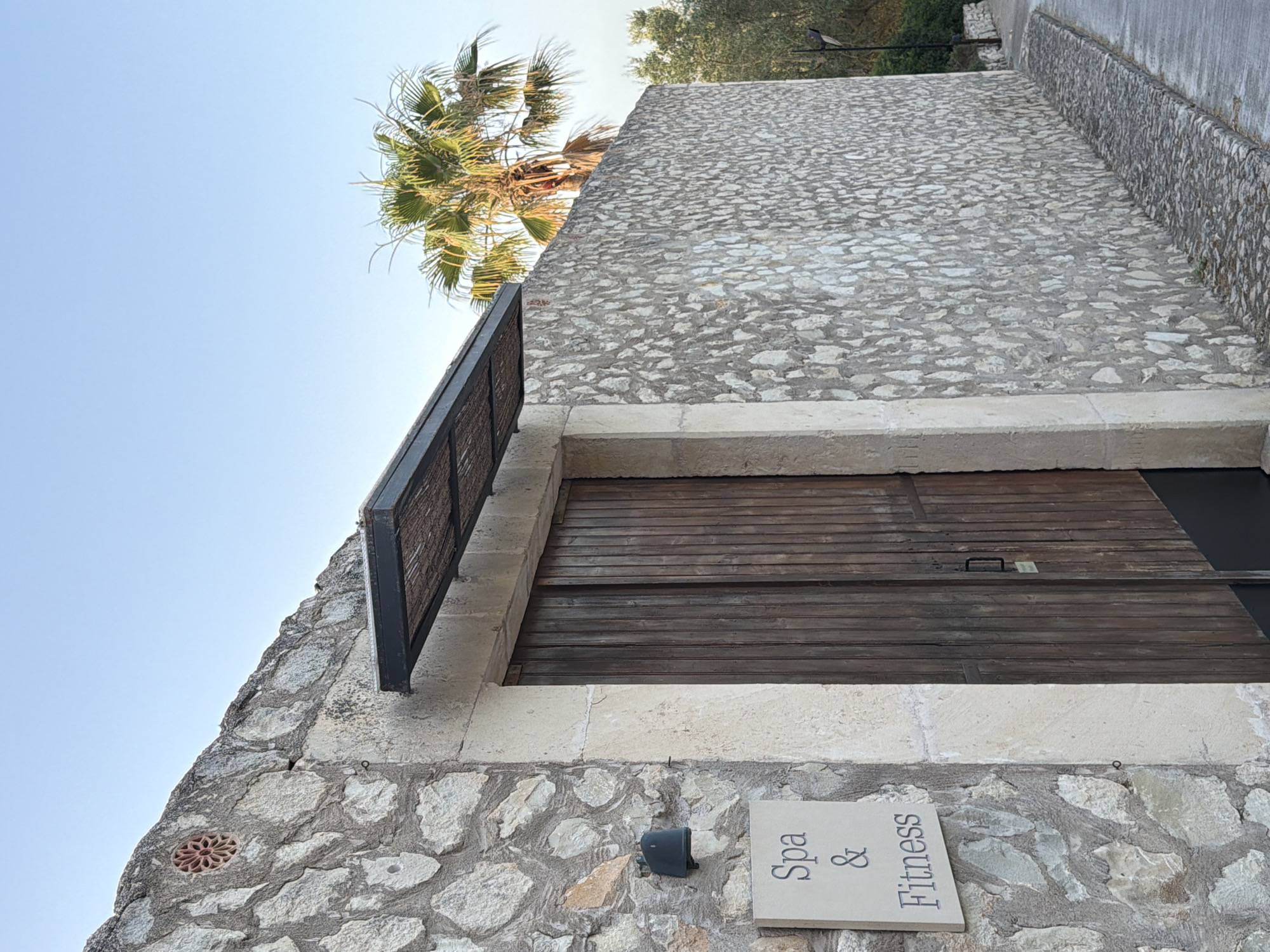 An exterior shot captures a rustic stone wall with a large wooden double door, likely an entrance. Above the door, a dark awning with a textured underside provides shade. To the left of the door, a stone plaque displays the words "Spa & Fitness." A black outdoor light fixture is mounted on the stone wall above the plaque. A palm tree is visible behind the stone wall and to the right of the door, with a clear blue sky overhead. The pathway in front of the entrance is paved with cobblestones. The scene appears to be in the town of Montuïri, Spain, given the architectural style and vegetation. There are no people or animals present.